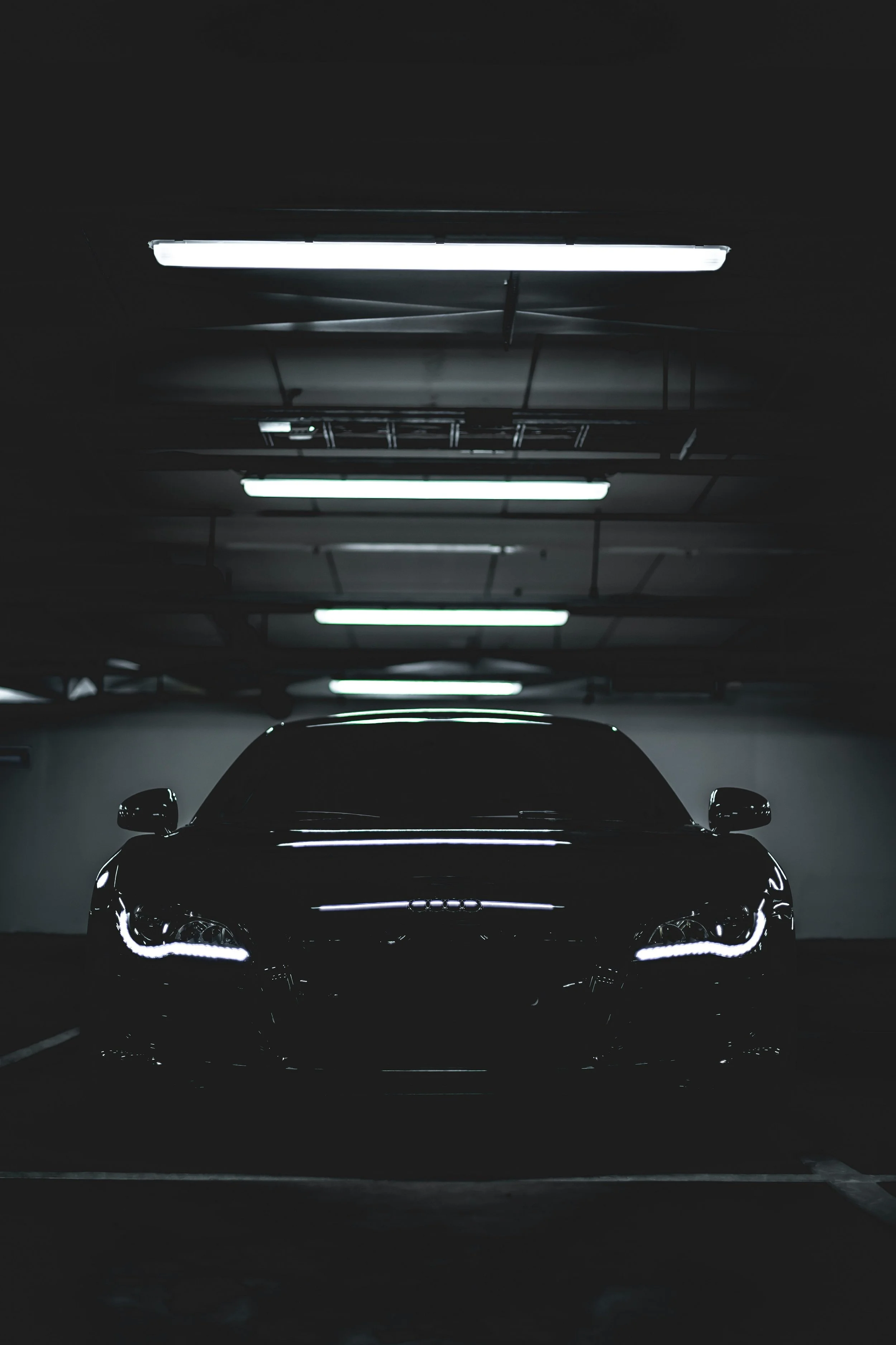 A black luxury car with LED headlights parked in a dimly lit underground garage with fluorescent overhead lights.