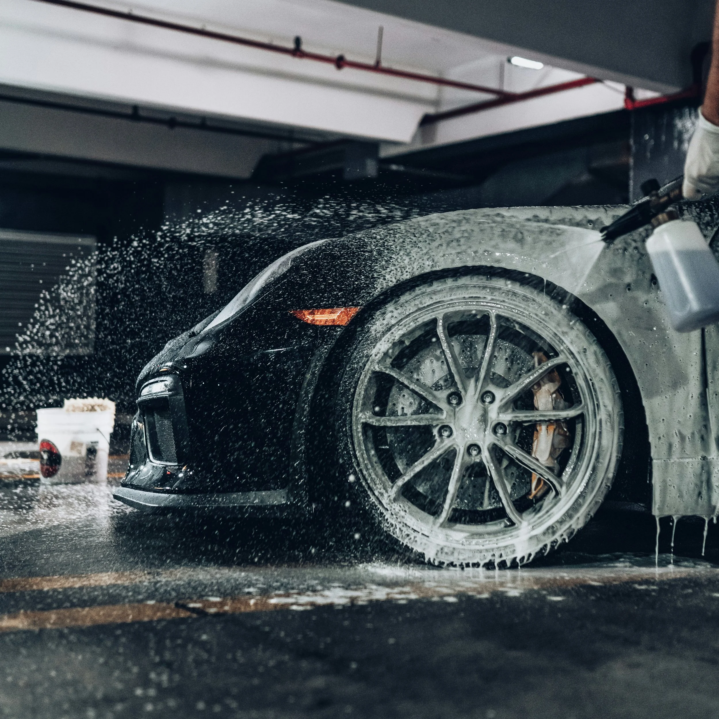 Close-up of a black car being washed in an automatic car wash, with soap and water splashing on the vehicle's tires and body.
