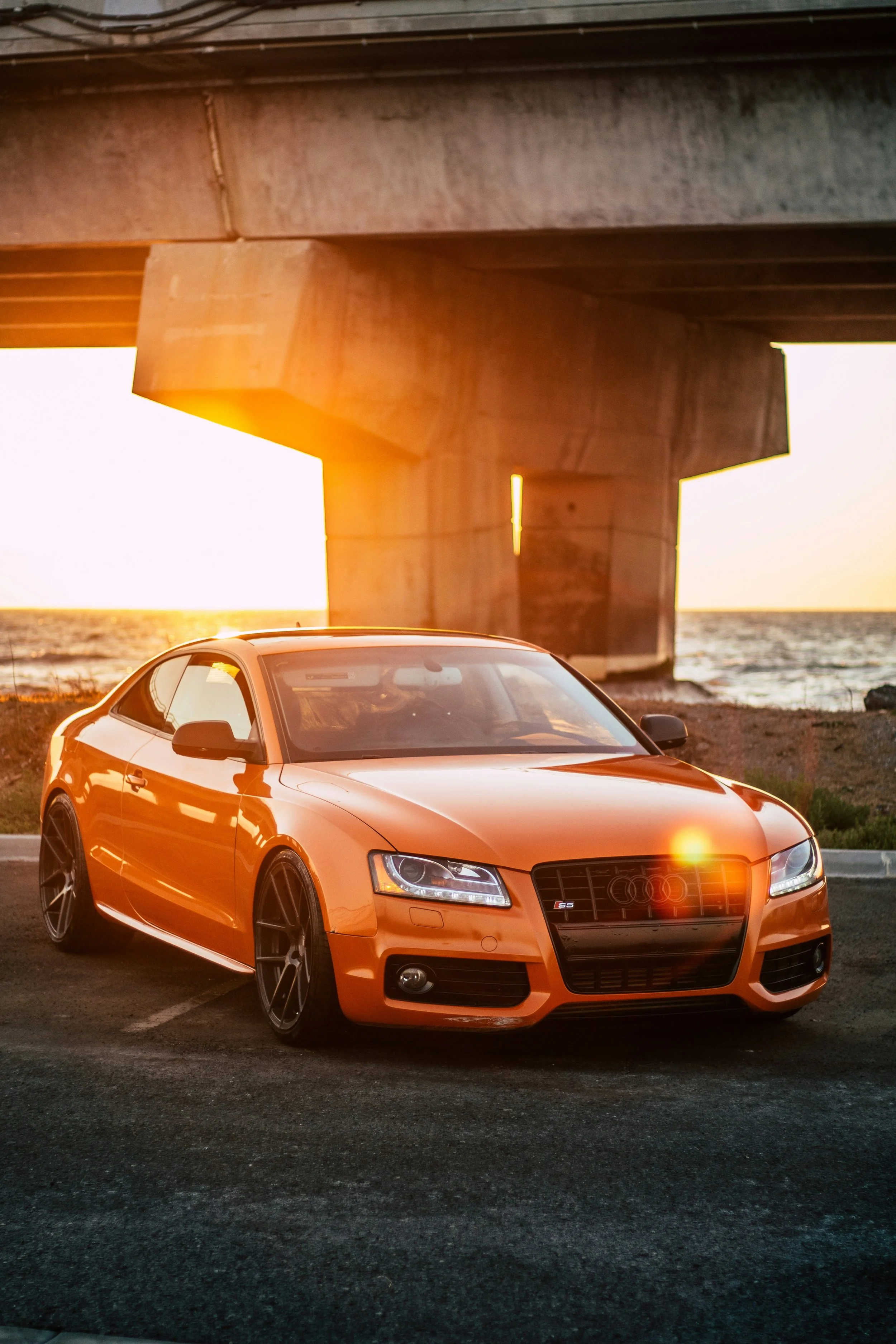 An orange Audi S5 parked on the side of a road near the coast during sunset, with the bridge in the background and the sun casting a warm glow.