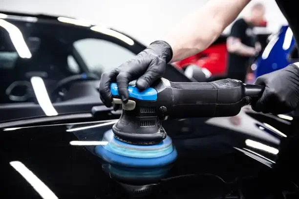 Person using a dual-action polisher with a black handle and blue backing pad on a black car's hood, with workshop environment in the background.