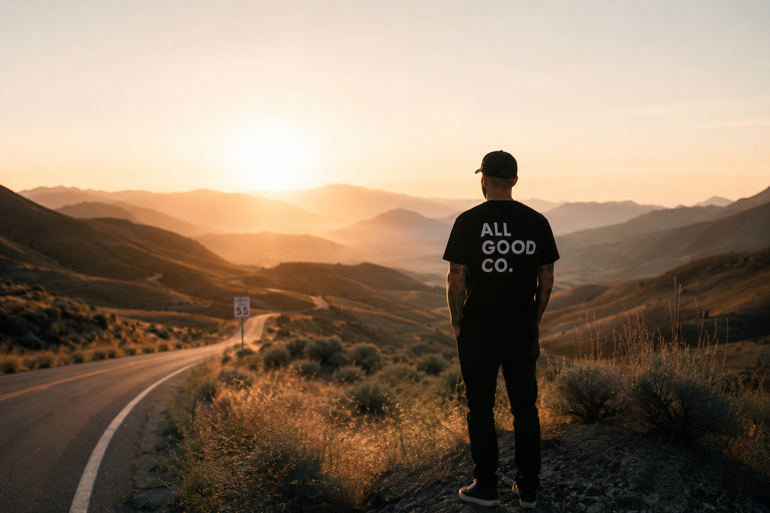 Man standing alone looking at the open road and mountains wearing a black hat and black t-shirt that says ALL GOOD CO.