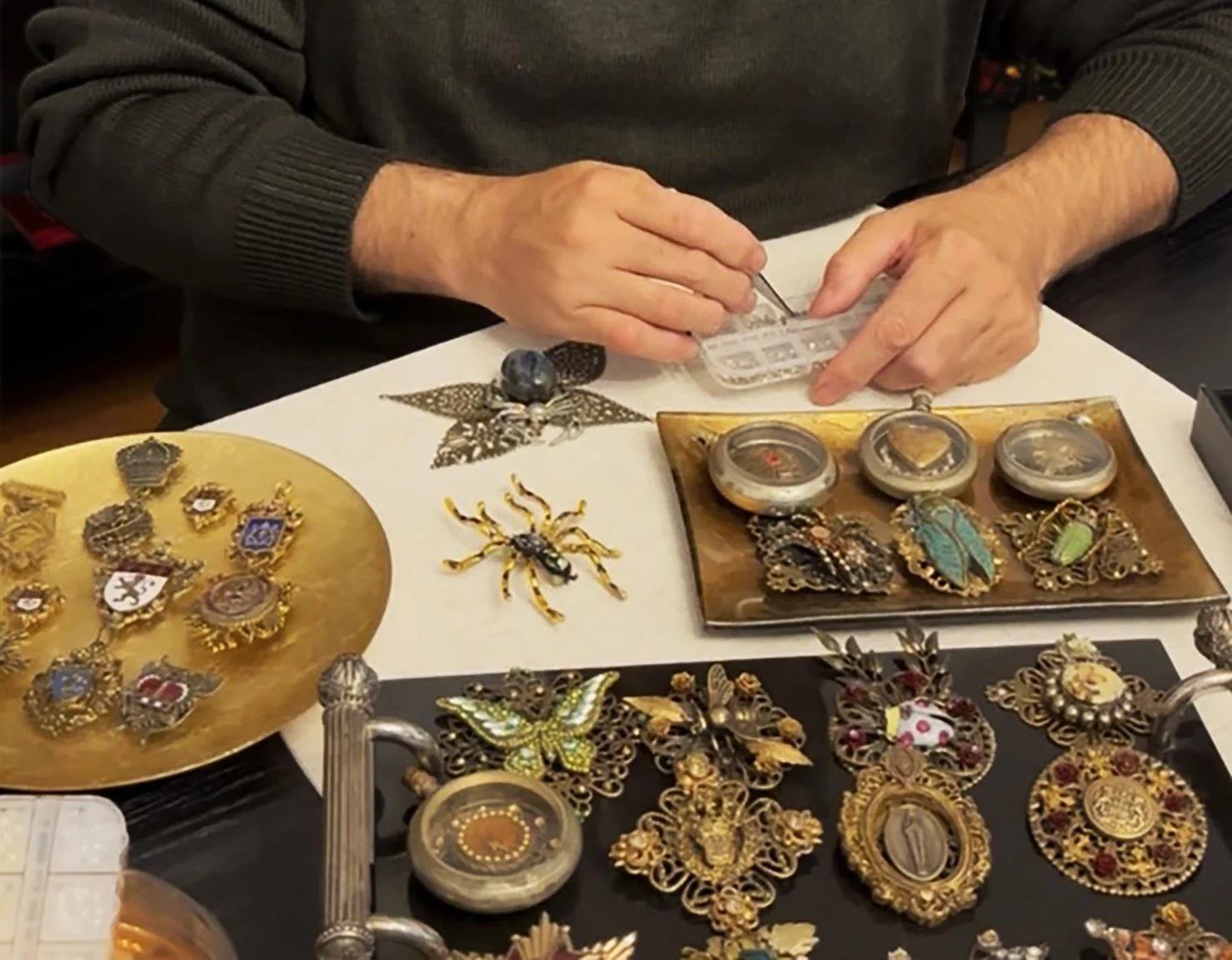 Person working with jewelry and collectibles, including brooches and pins, on a table with various decorative items.