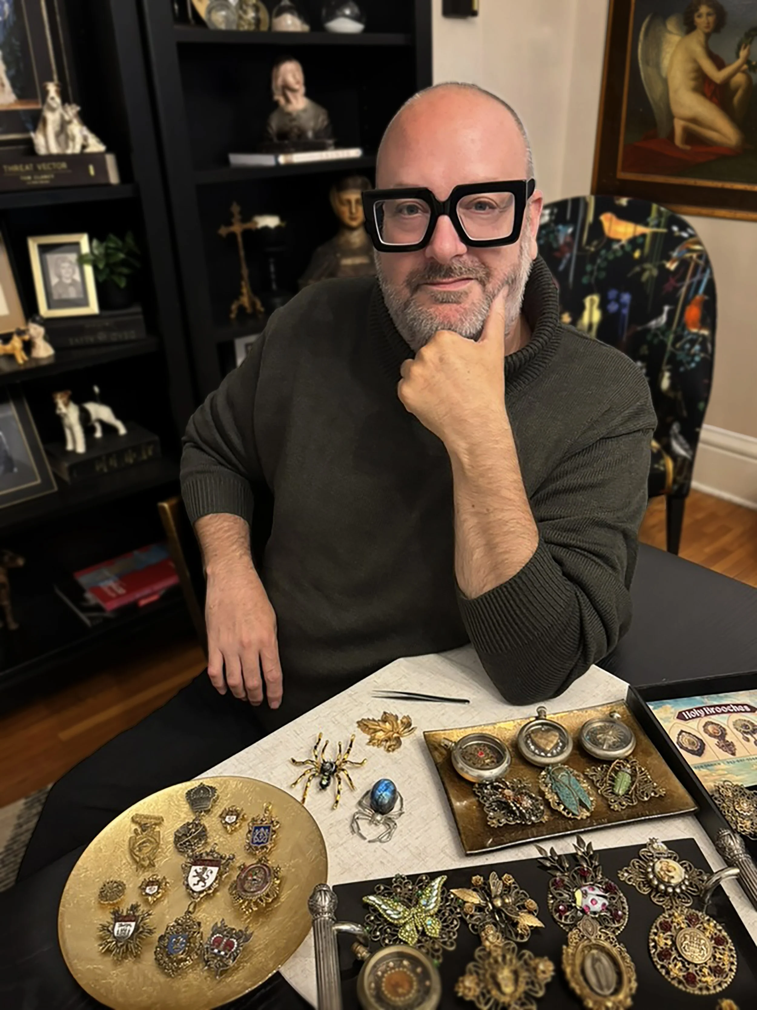 A man wearing oversized black glasses sitting at a table with various vintage jewelry, jewelry boxes, and collectible items, with books and framed photos on shelves and artwork on the wall in the background.