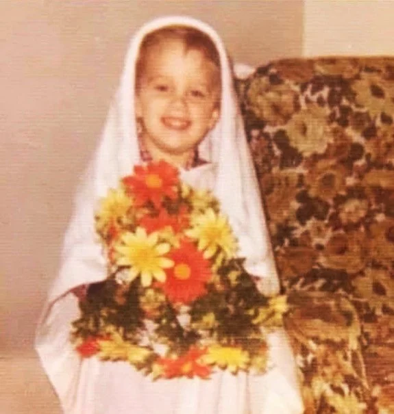 Photograph of a young boy dressed in white, wearing a cloth veil, holding a large bouquet of colorful flowers, sitting on a patterned armchair.