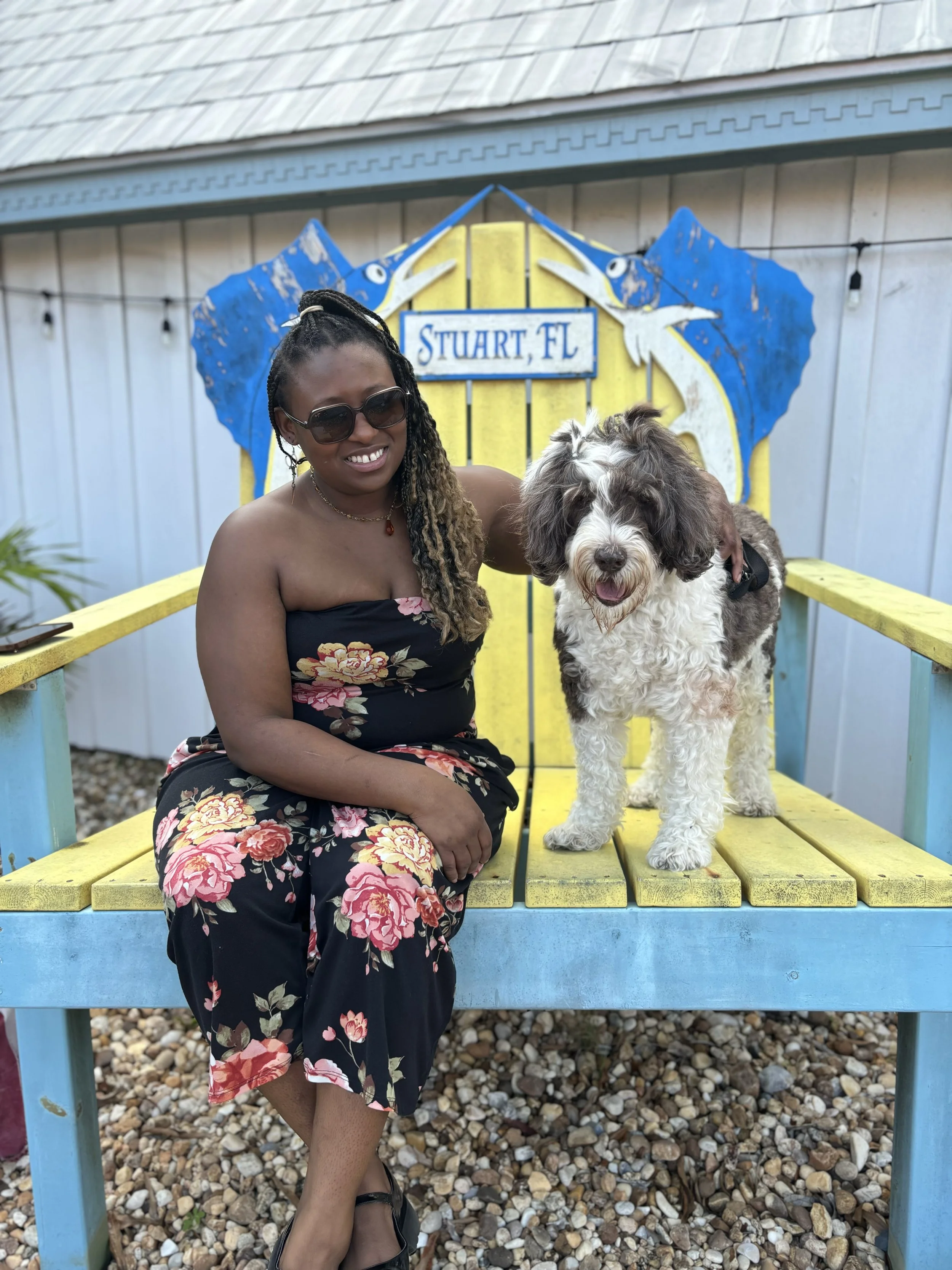 A woman sitting on a yellow and blue bench with a fluffy dog beside her near a building with a sign that reads 'Stuart, FL' and a blue, white, and yellow mural of two marlins.