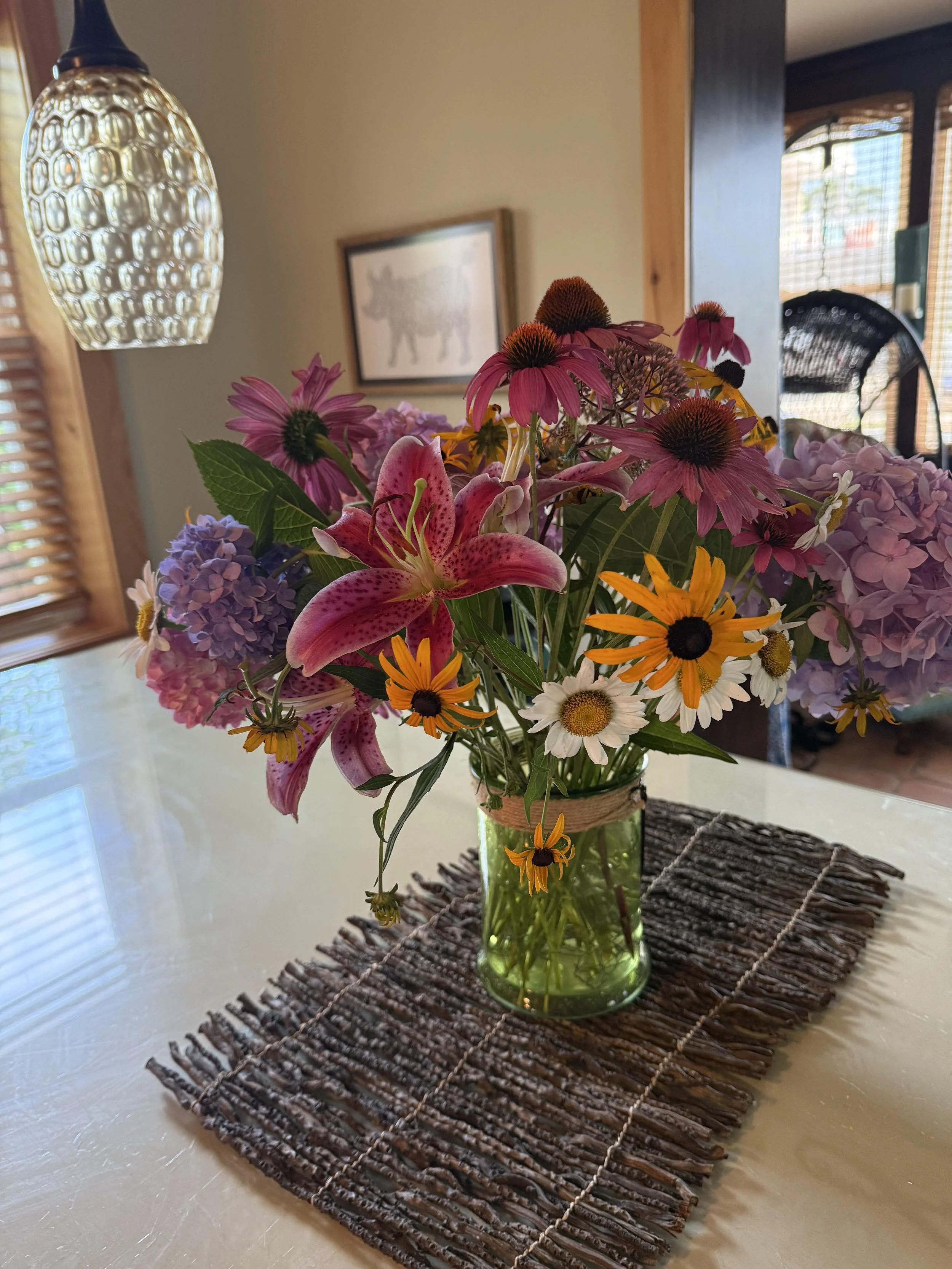 A glass vase with a bouquet of pink lilies, purple hydrangeas, orange and yellow coneflowers, and other mixed flowers on a white table. The vase is placed on a dark woven placemat. In the background, there is a framed picture of a dog on the wall, a hanging lamp, and a room with windows and wooden accents.