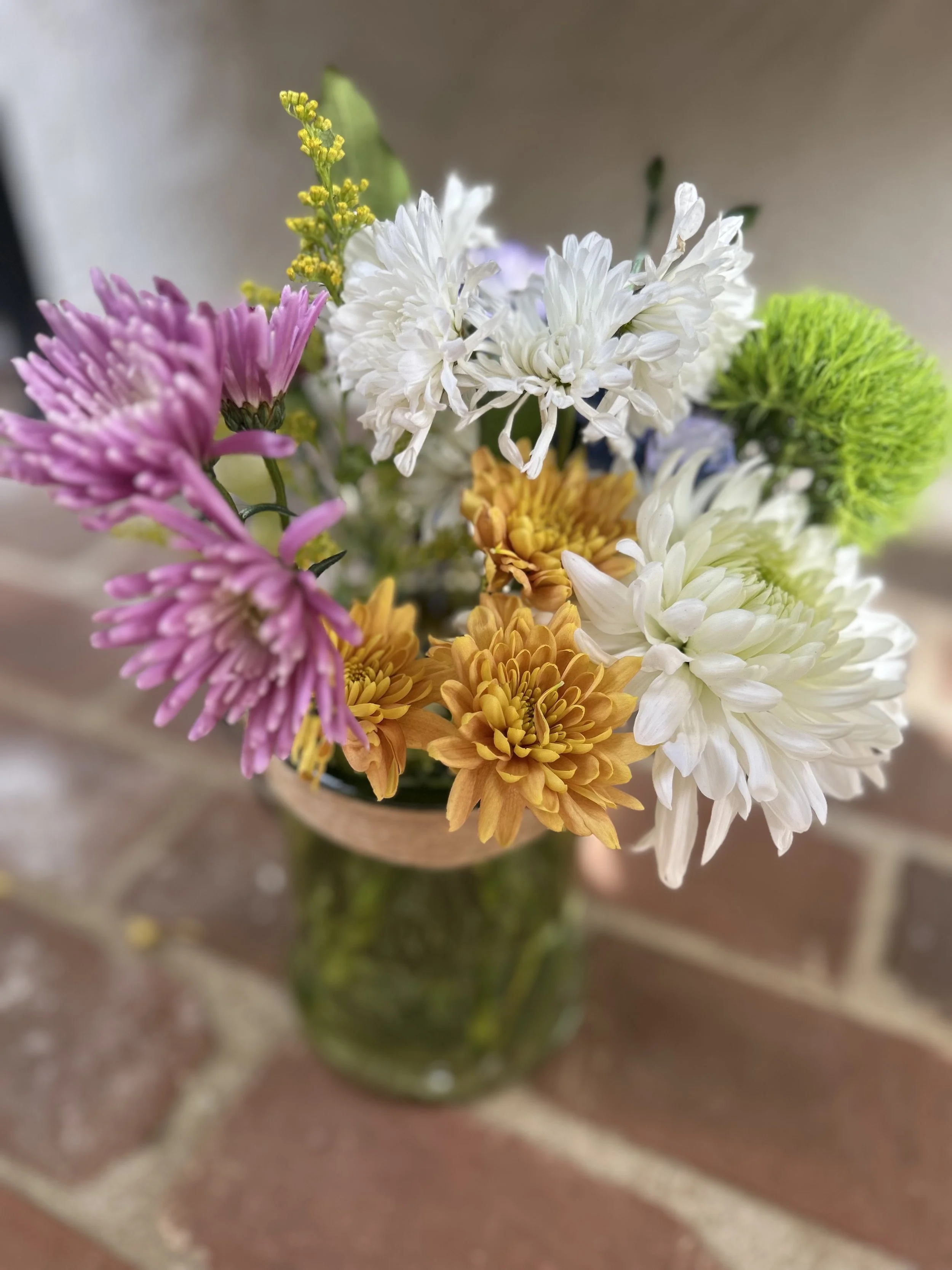 Close-up of a bouquet of colorful flowers in a green vase on a brick surface.