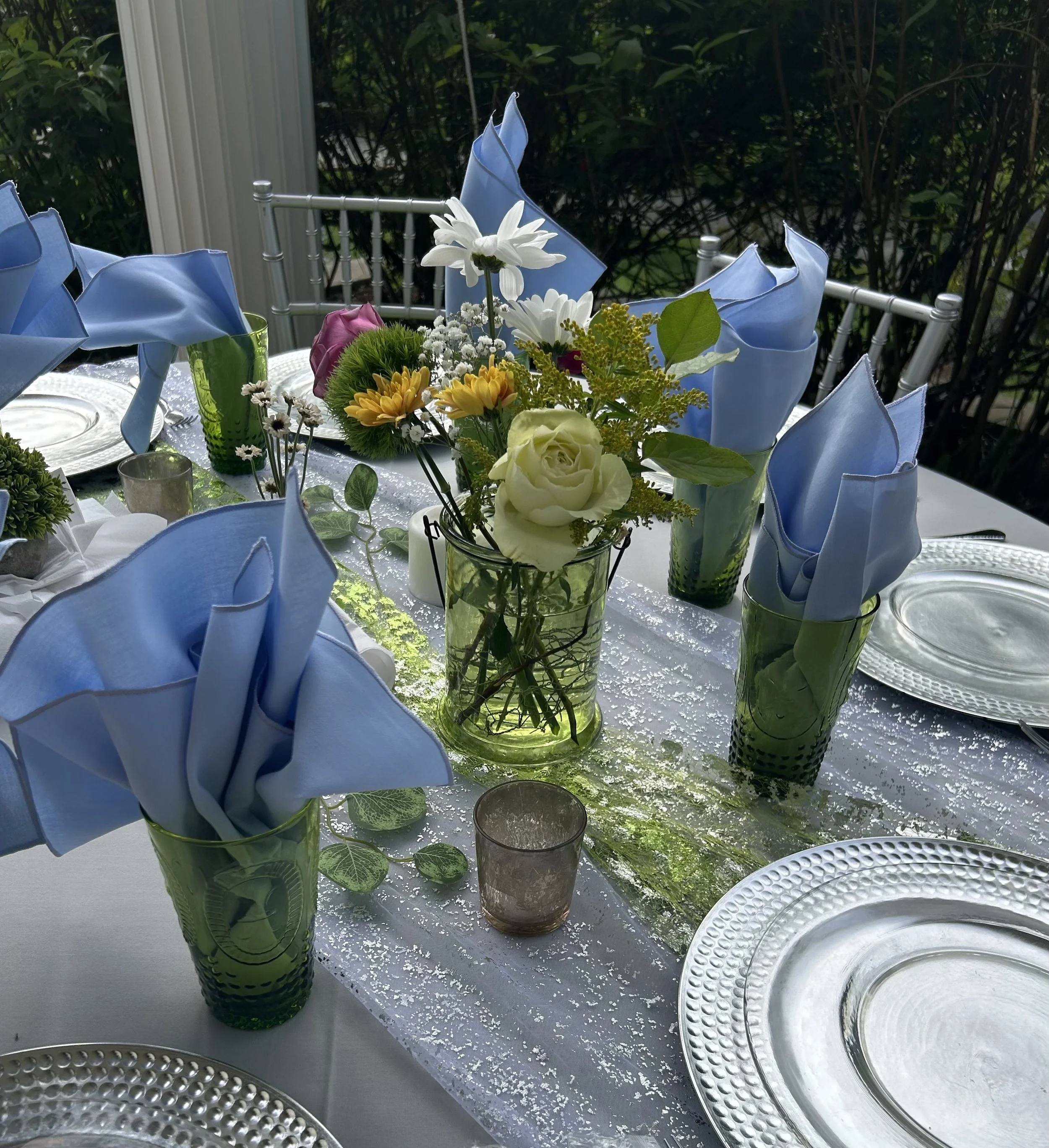 A table set for a meal with a floral centerpiece and blue napkins in green glasses, along with white plates and silverware, and surrounded by silver chairs, outdoors with greenery in the background.