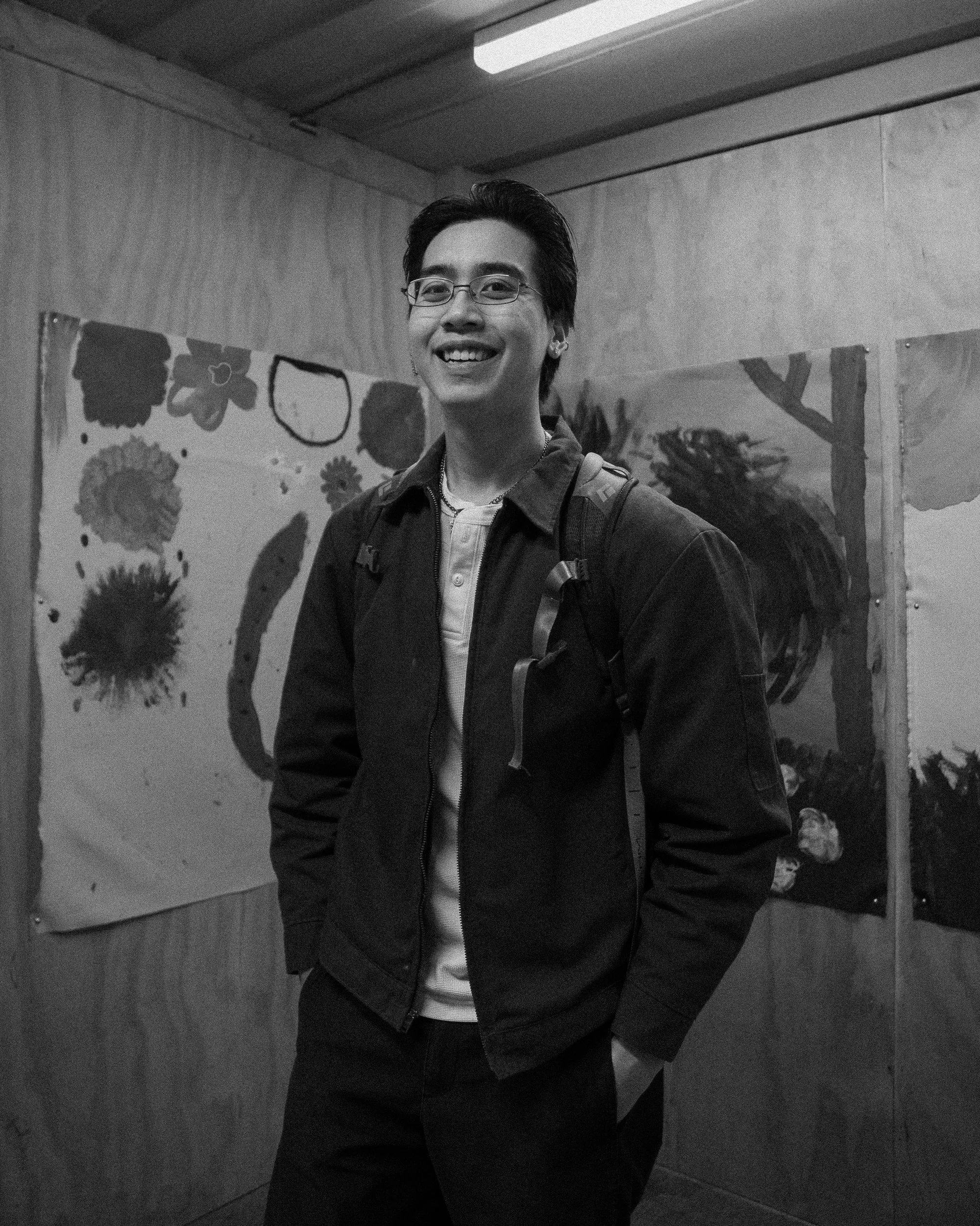 A young man with short dark hair, glasses, and earrings smiling while standing indoors in front of a wall decorated with artwork including flowers and a tree. He is wearing a dark jacket over a shirt and has a backpack strap on his shoulder.