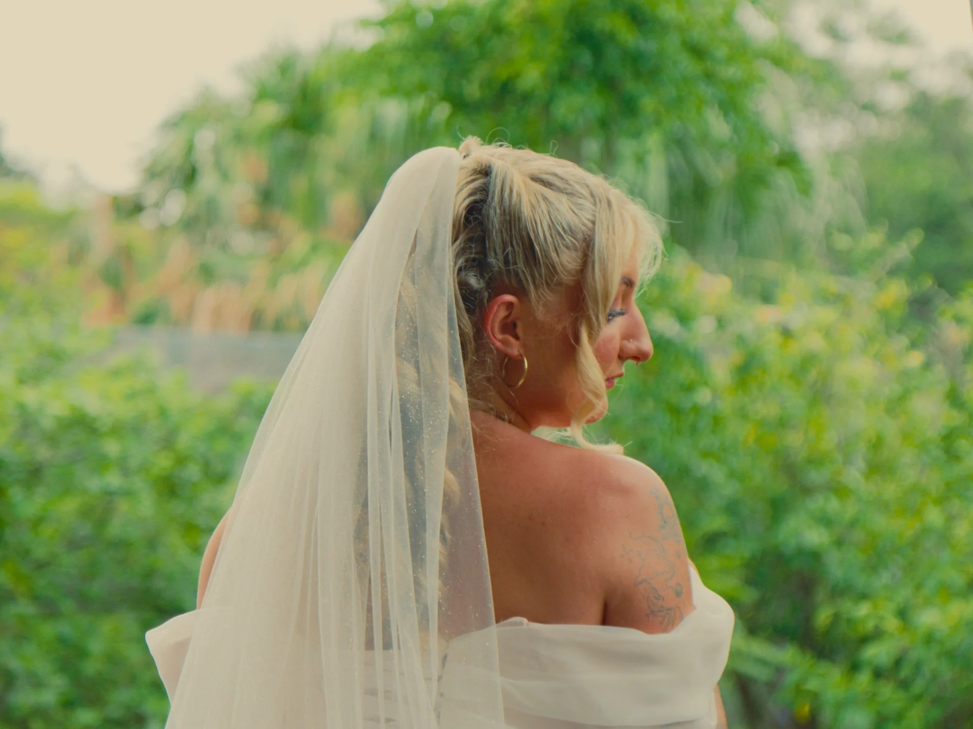 A woman with short blonde hair and tattoos on her arm is wearing a white strapless dress and a light veil, standing outdoors with a blurred green foliage background.