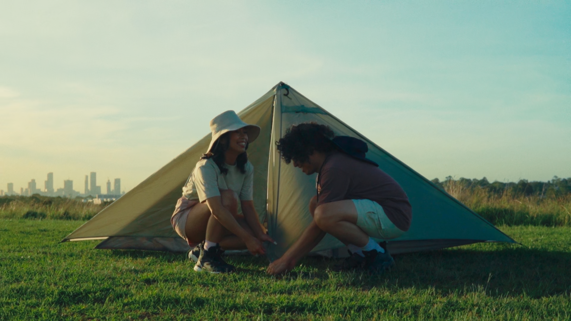 Two young adults, a woman and a man, are setting up a small tent on a grassy field during sunset with a city skyline visible in the background. The woman wears a wide-brimmed hat and shorts, smiling at the man who is crouched down securing the tent, dressed in casual gear.