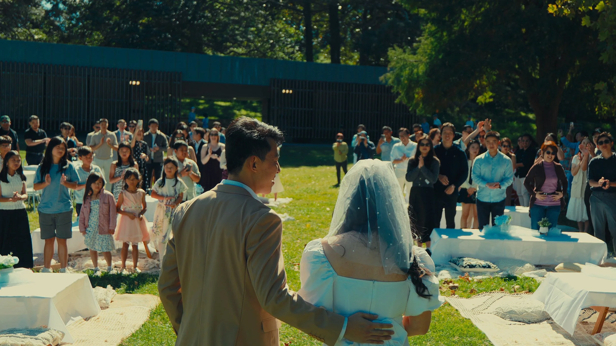 Outdoor wedding ceremony with a crowd of guests, bride and groom, and tables with cakes and decorations in a park with green trees.