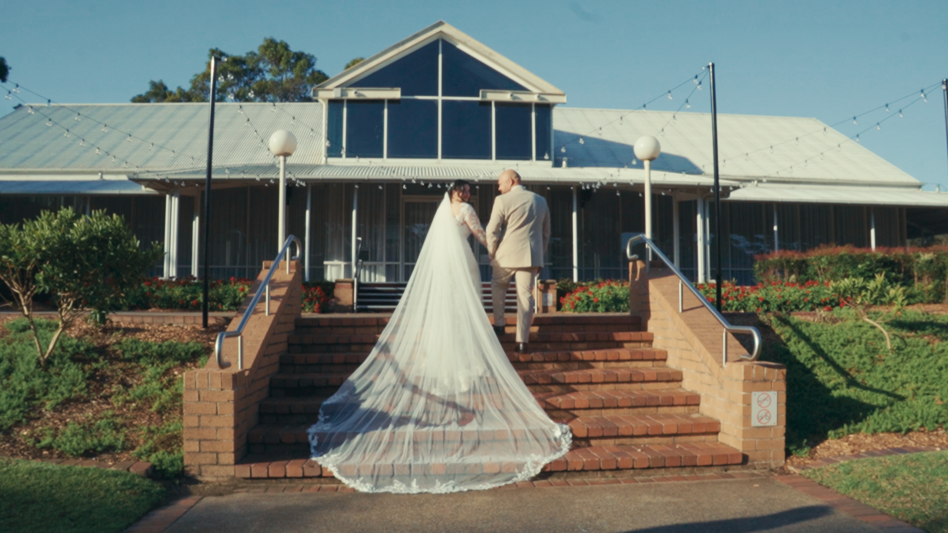Bride and groom holding hands on the steps of a building with string lights above, greenery on side, and a blue sky.