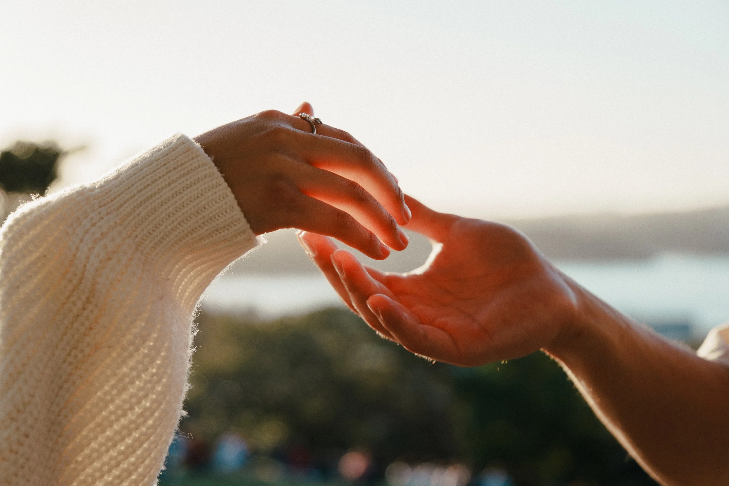 Two hands reaching towards each other against a blurred outdoor background, one hand appears to be giving and the other receiving a gentle touch.