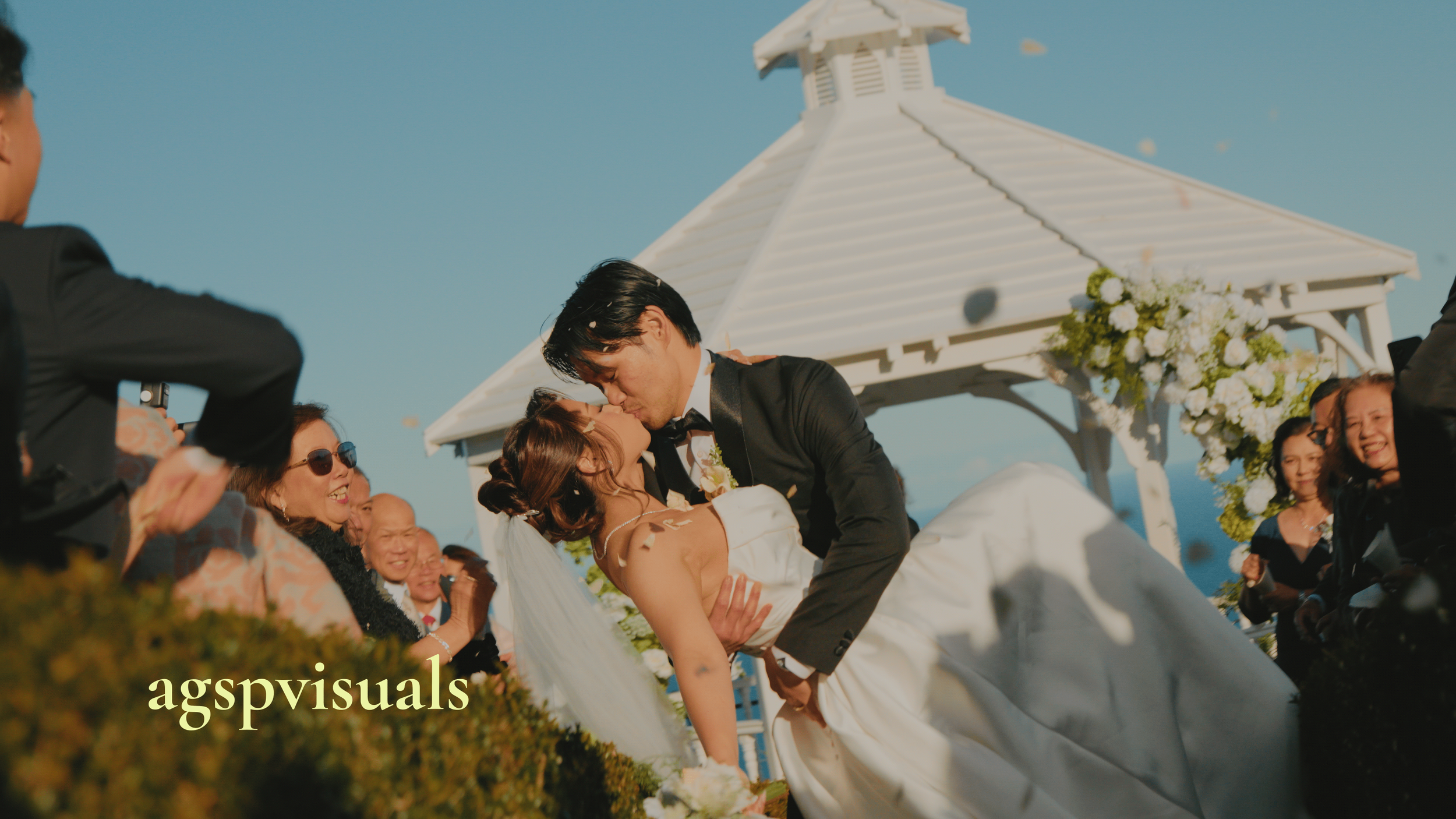 A bride and groom share a kiss at their outdoor wedding ceremony, surrounded by friends and family, under a white gazebo with floral decorations, during daytime with a clear blue sky.