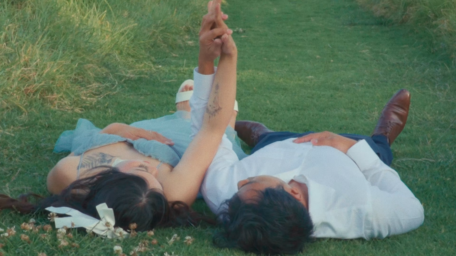 Two people lying on the grass, with one touching the other's hand in a courting gesture.