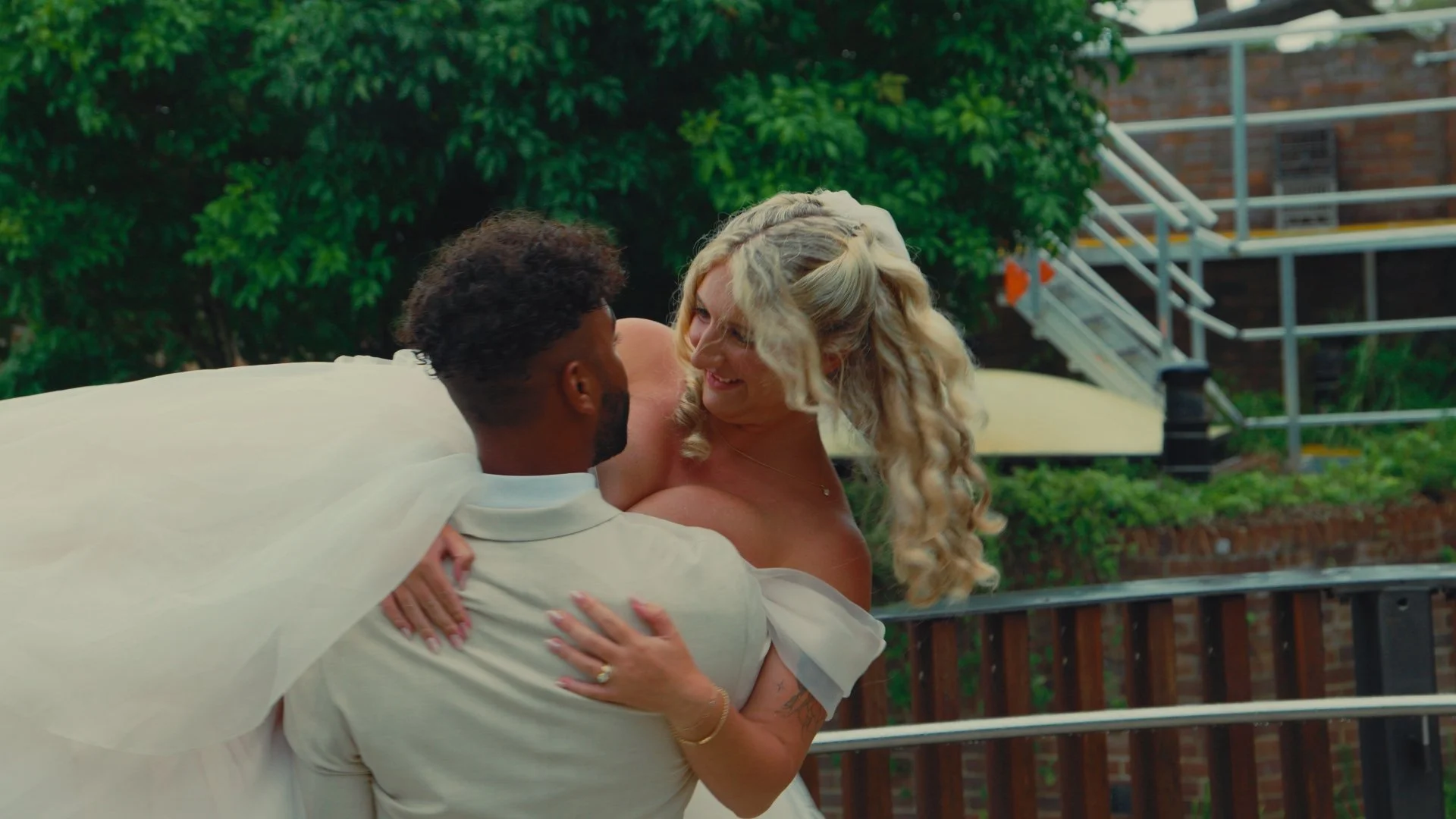 A man in a white shirt is holding a smiling woman with long blond curly hair, wearing a white dress, in an outdoor setting with a green leafy background and a brick building with metal stairs.