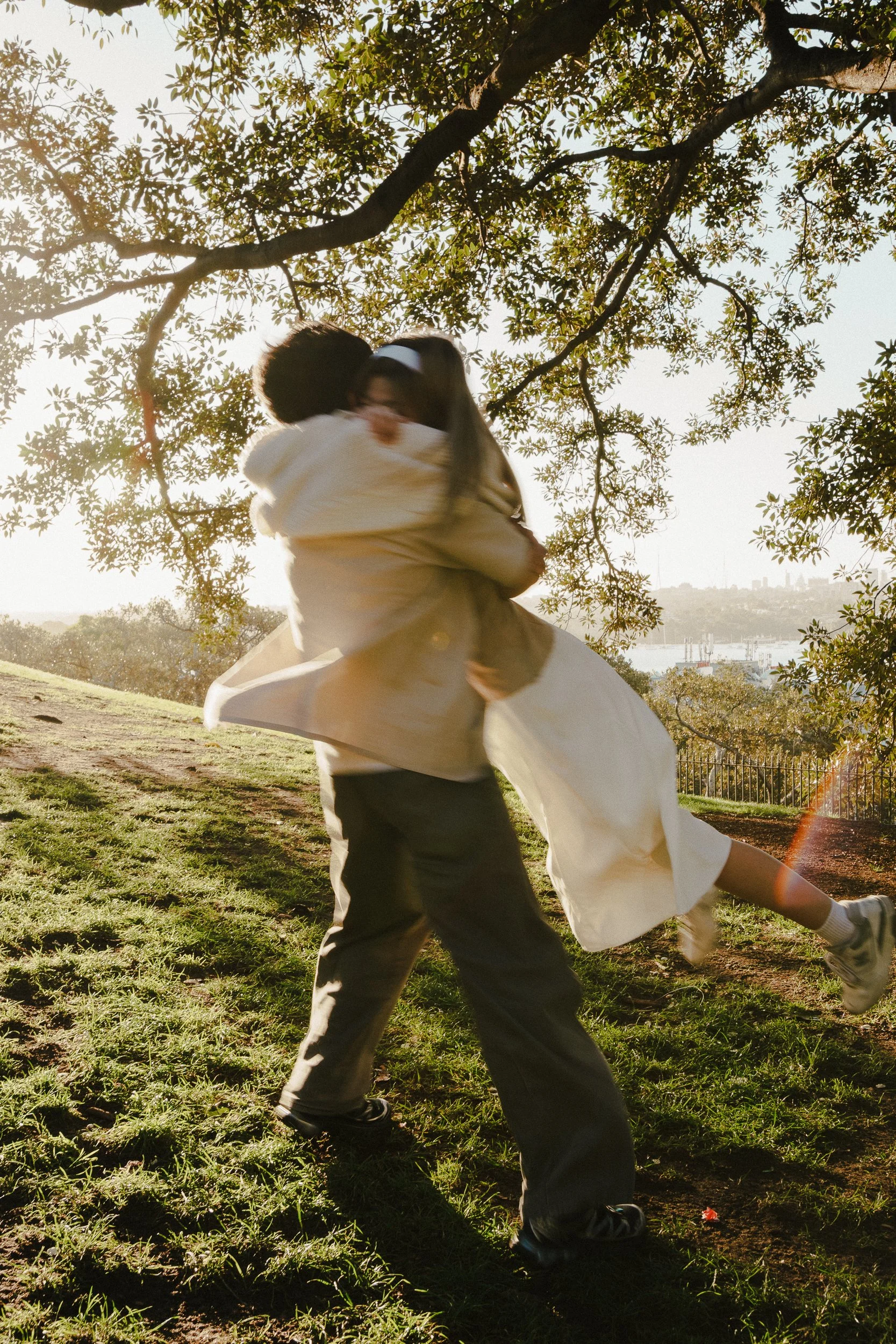 Two people dancing outdoors on a grassy area near a tree, with the sun shining through the leaves and water in the background.