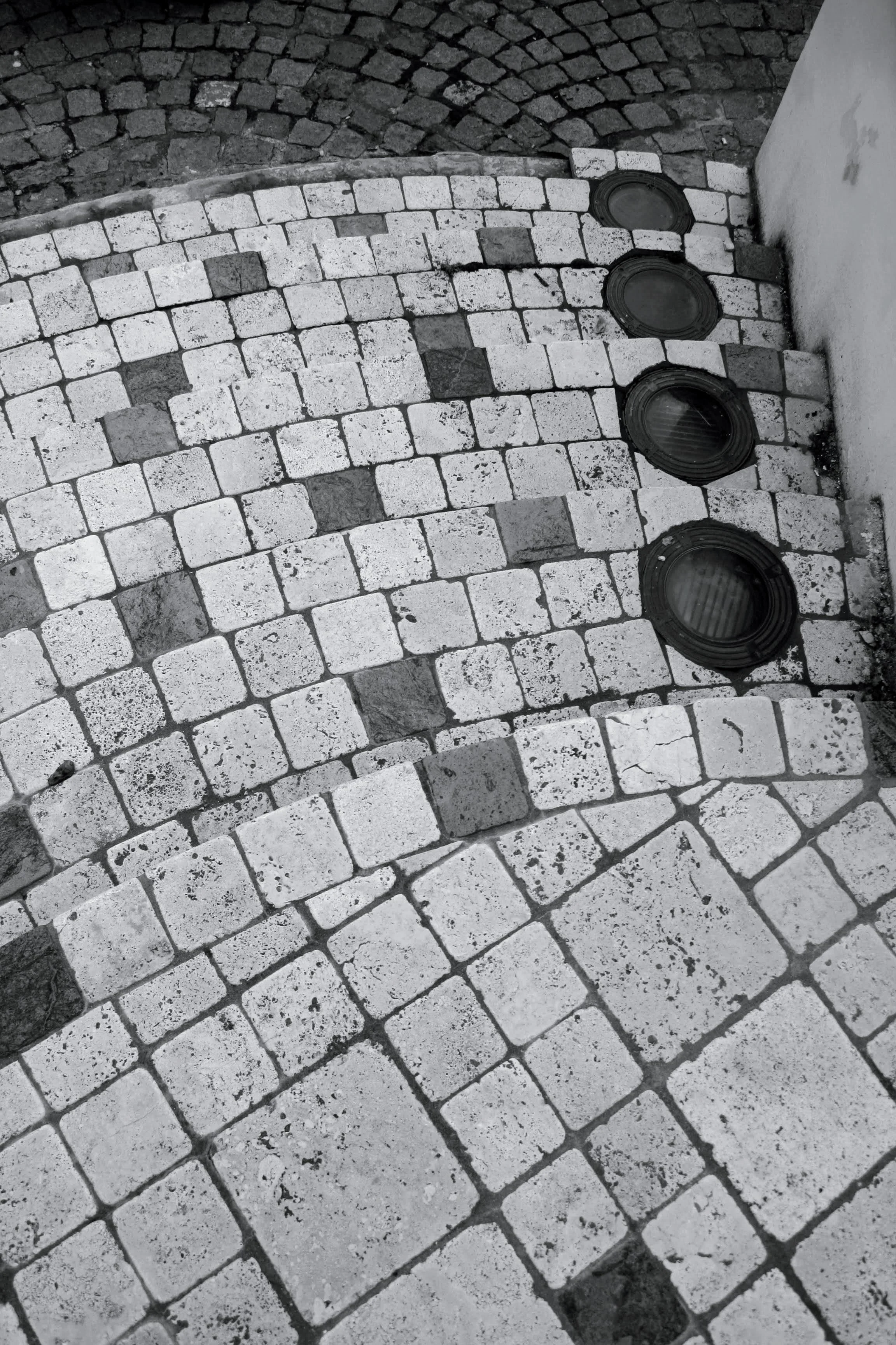 Pavés ronds anciens en noir et blanc, avec des bouches d'égout circulaires le long d'un mur.