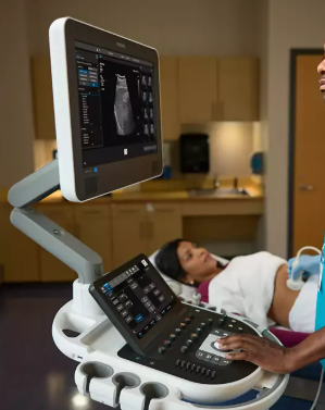 Ultrasound machine displaying an ultrasound scan with a patient lying on a bed nearby in a medical examination room.