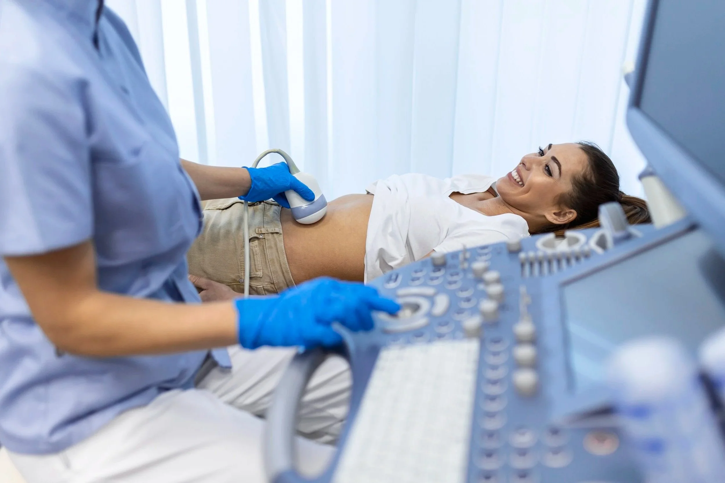 A woman lying on an ultrasound examination table, smiling, during a medical ultrasound procedure performed by a healthcare professional.