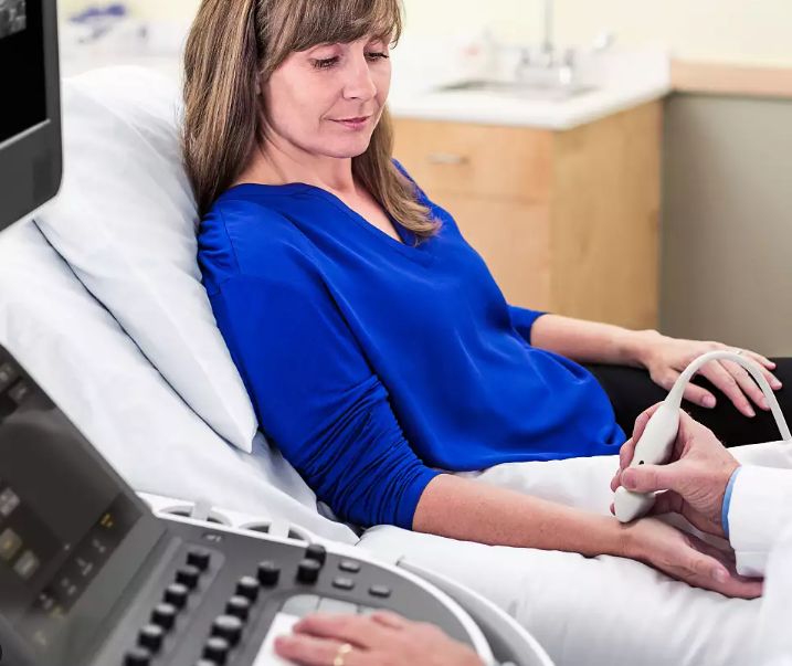 A woman in a blue shirt lying on a hospital bed while a healthcare professional conducts an ultrasound on her forearm.