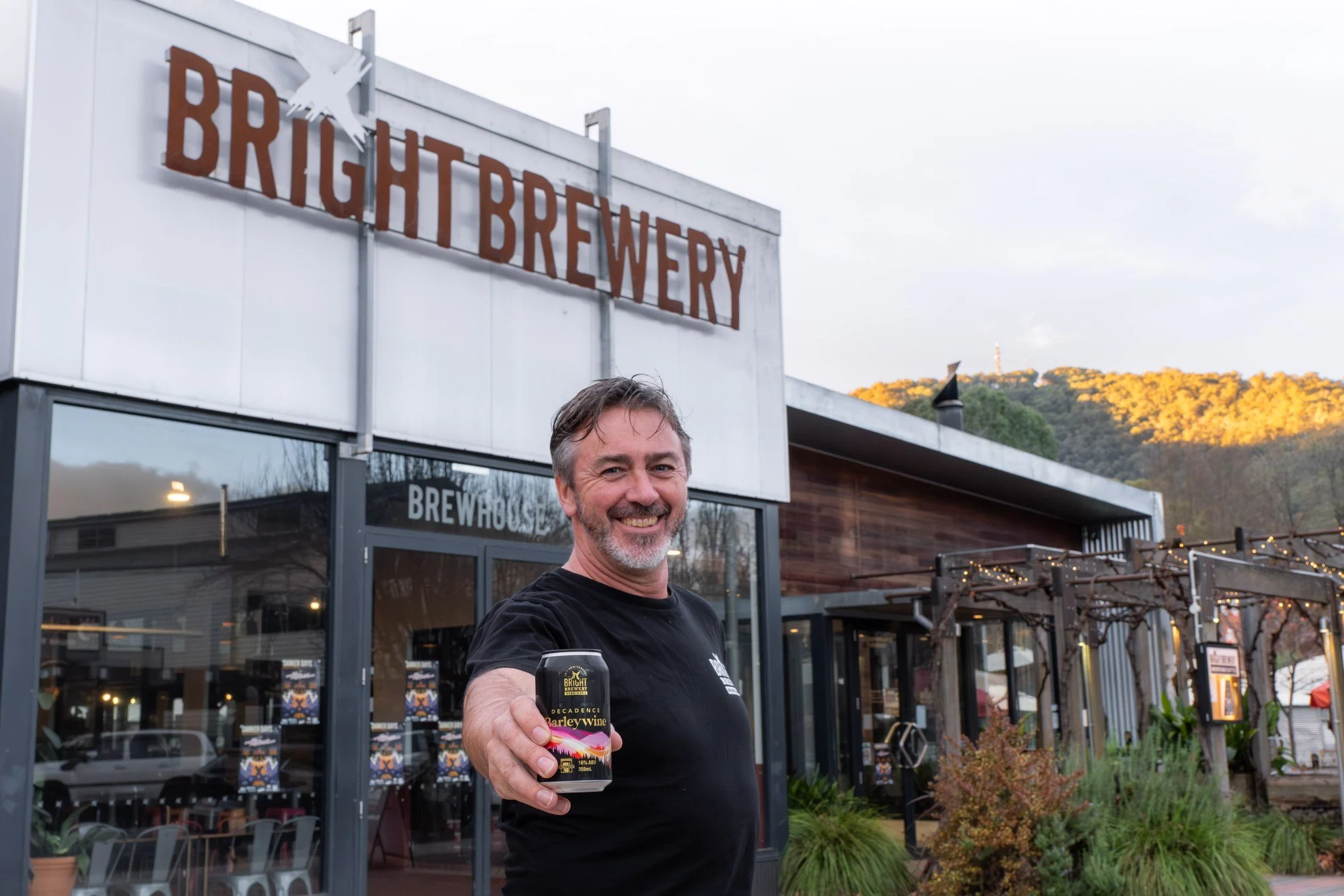 A man smiling and holding a can of Bright Brewery beer outside of Bright Brewery in a mountain town, with a sign reading 'Bright Brewery' and a background of trees and hills.