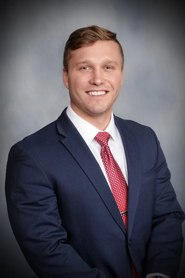 Portrait of a young man in a dark blue suit, white shirt, and red patterned tie, smiling against a gray background.