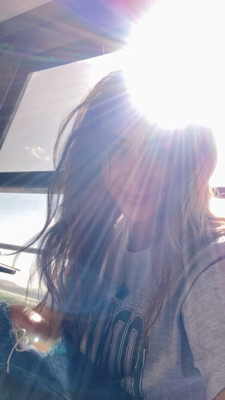 A person with long hair is sitting by a window with sunlight shining directly on them, creating lens flare and partially obscuring their face.
