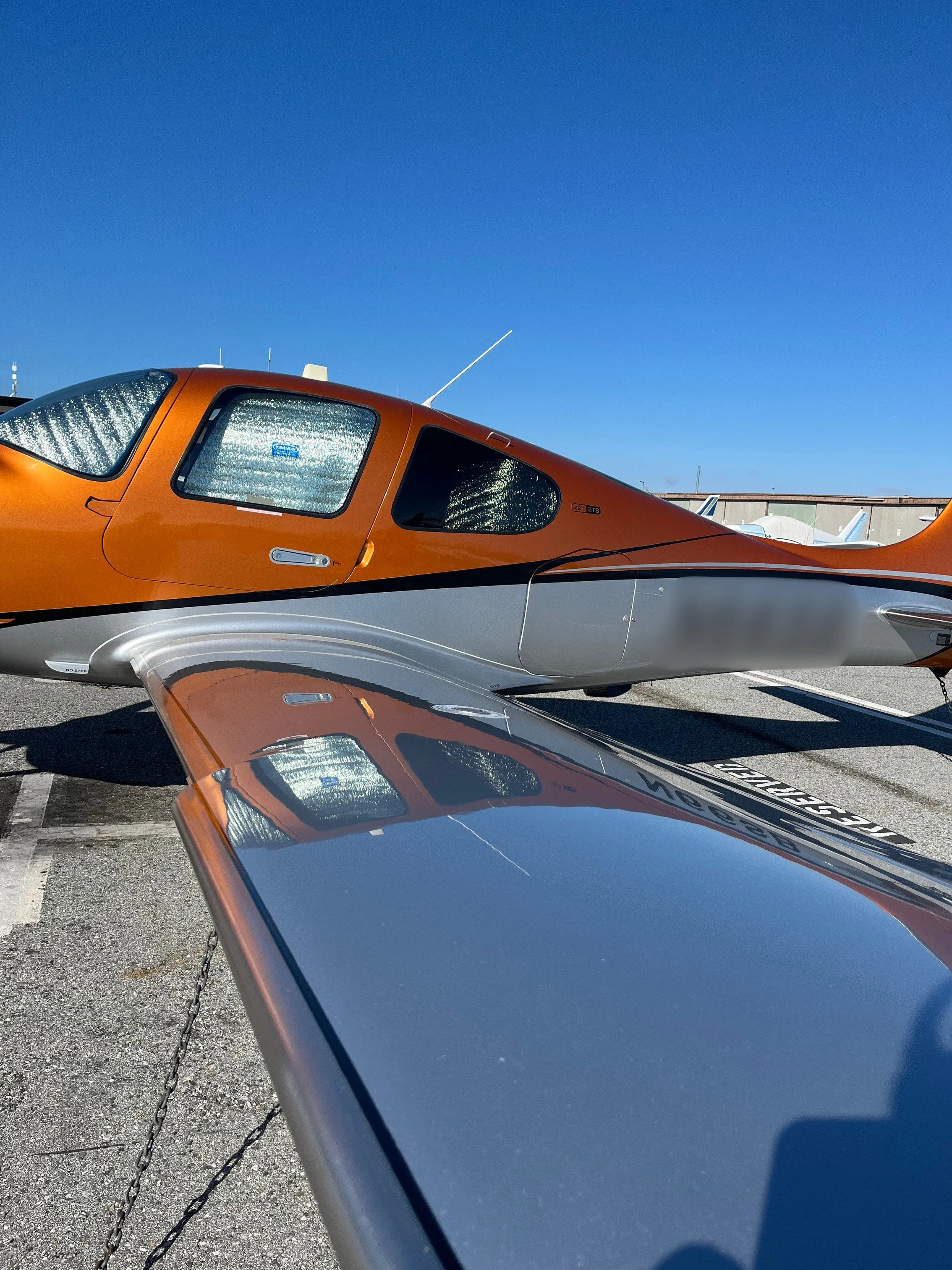 Close-up of a small orange and gray airplane parked on an airport tarmac, with other aircraft and hangars in the background as the sky is clear and blue.