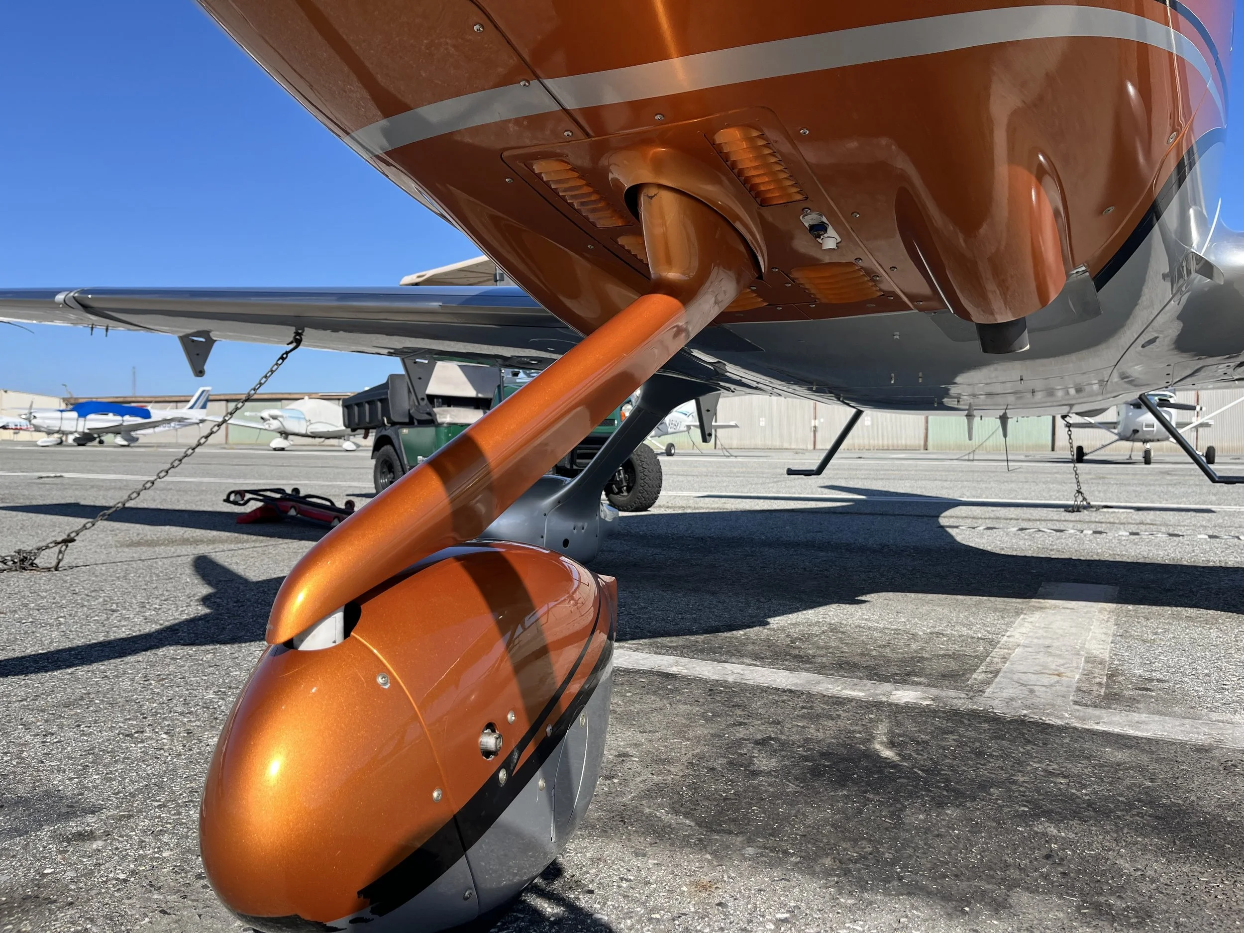 Close-up of the underside of an orange small aircraft or glider, showing the landing gear and parts of the tail and wings. In the background, other small aircraft are visible at an airfield under a clear blue sky.