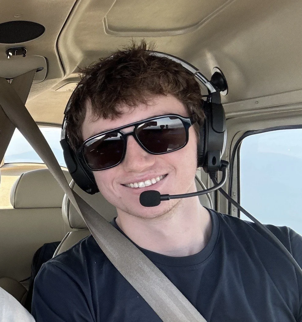 Young man smiling with sunglasses and wearing a headset with microphone, sitting in the cockpit of a small aircraft.