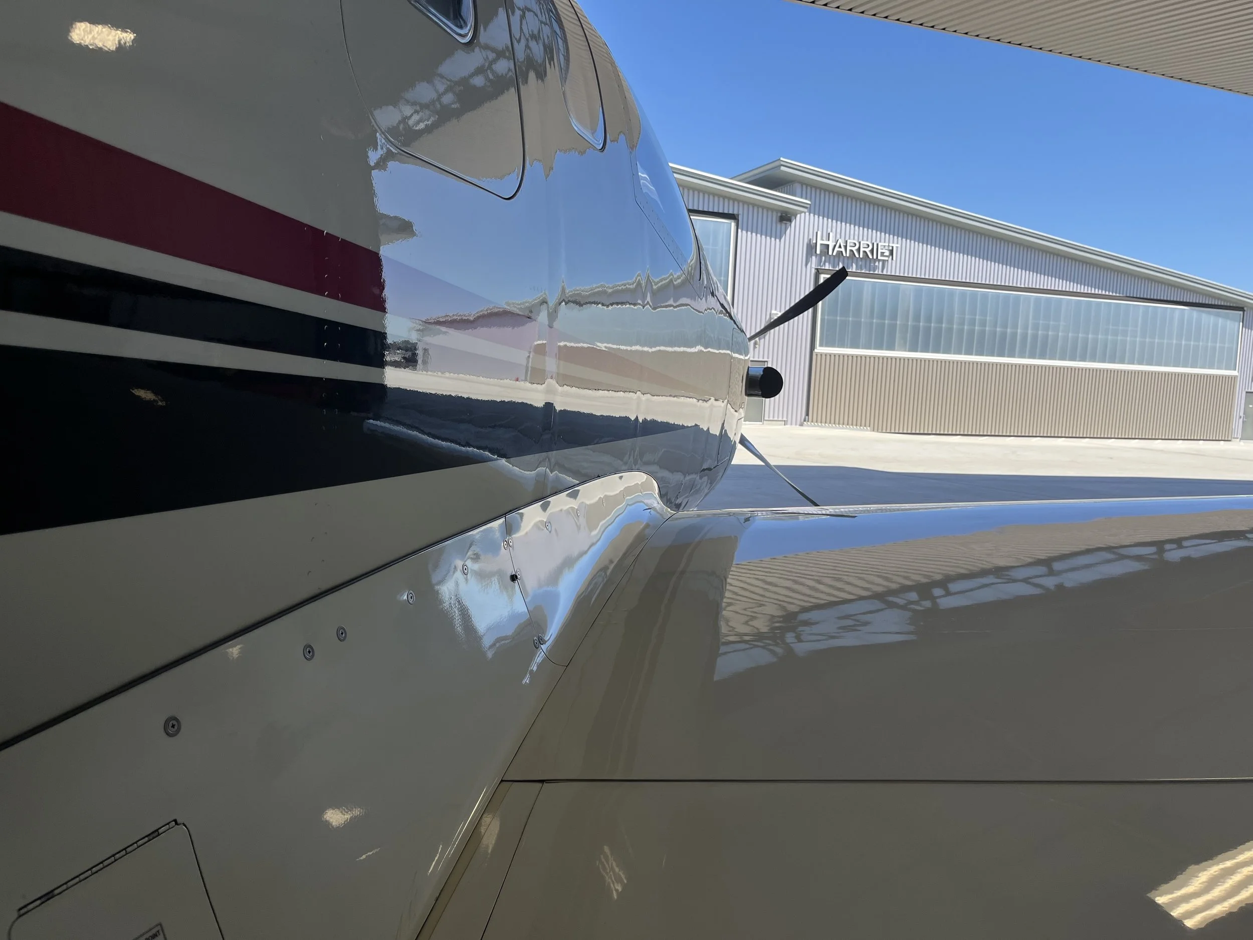 Close-up of a sleek, shiny aircraft with metallic surface, parked outside a modern building with the sign 'HARRIET' under a clear blue sky.