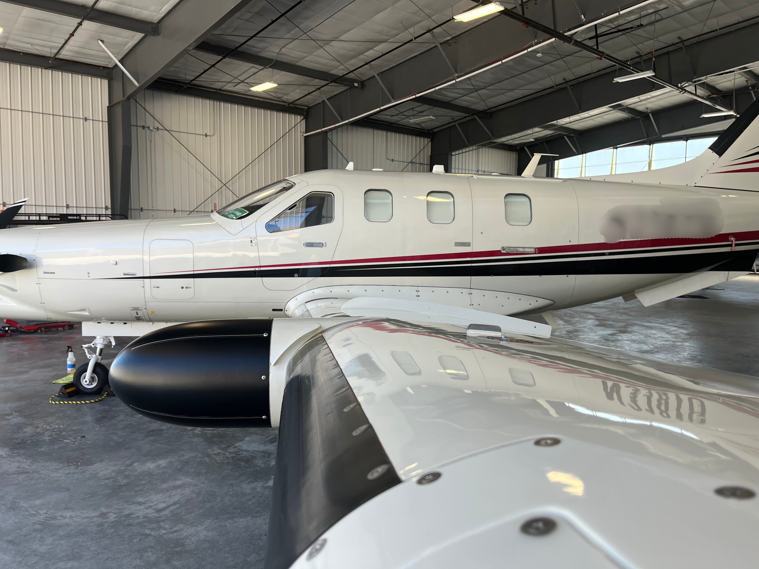 Inside a hangar, a white private jet with red and black accents is parked. The image shows part of the jet's fuselage, wing, and engine, with the hangar's ceiling and walls in the background.