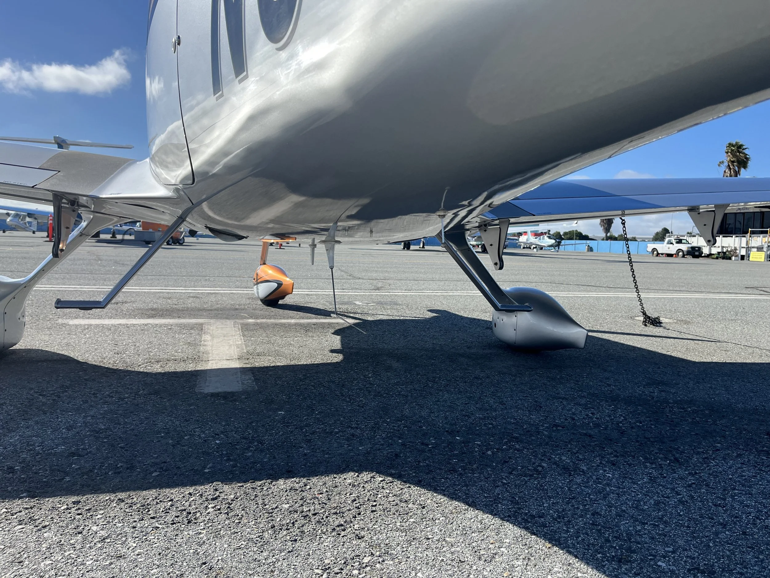 A small aircraft parked on an airport tarmac, with other planes and airport vehicles in the background, and a palm tree visible against a blue sky.