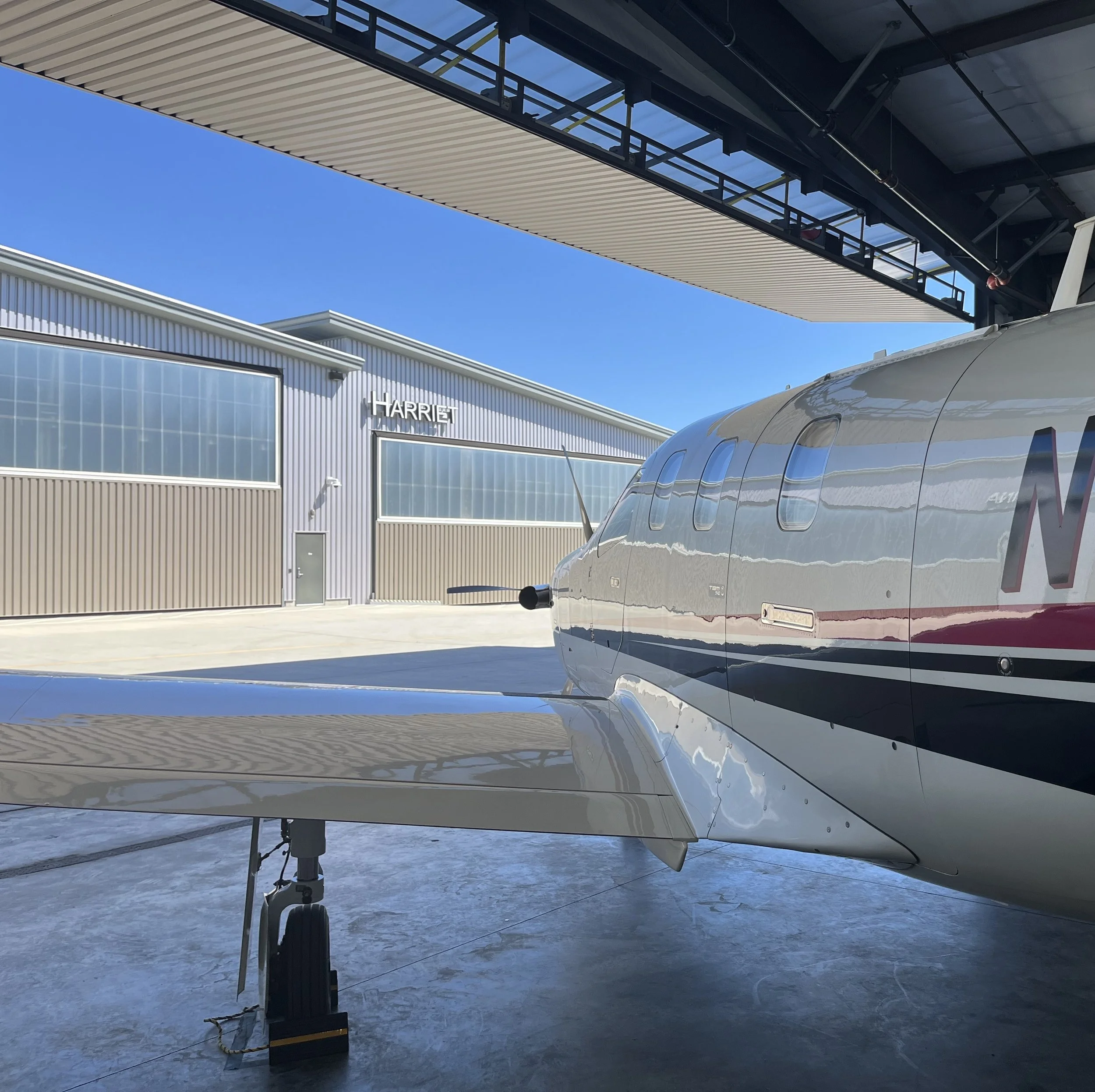 View of a private jet parked inside a hangar with a building labeled 'HARRIET' in the background, under a clear blue sky.
