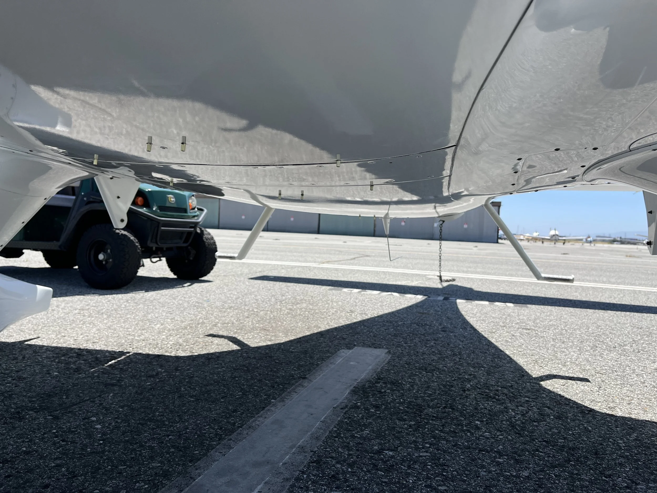 Close-up of the underside of a small aircraft wing on a tarmac, with part of a golf cart and other planes visible in the background under clear skies.