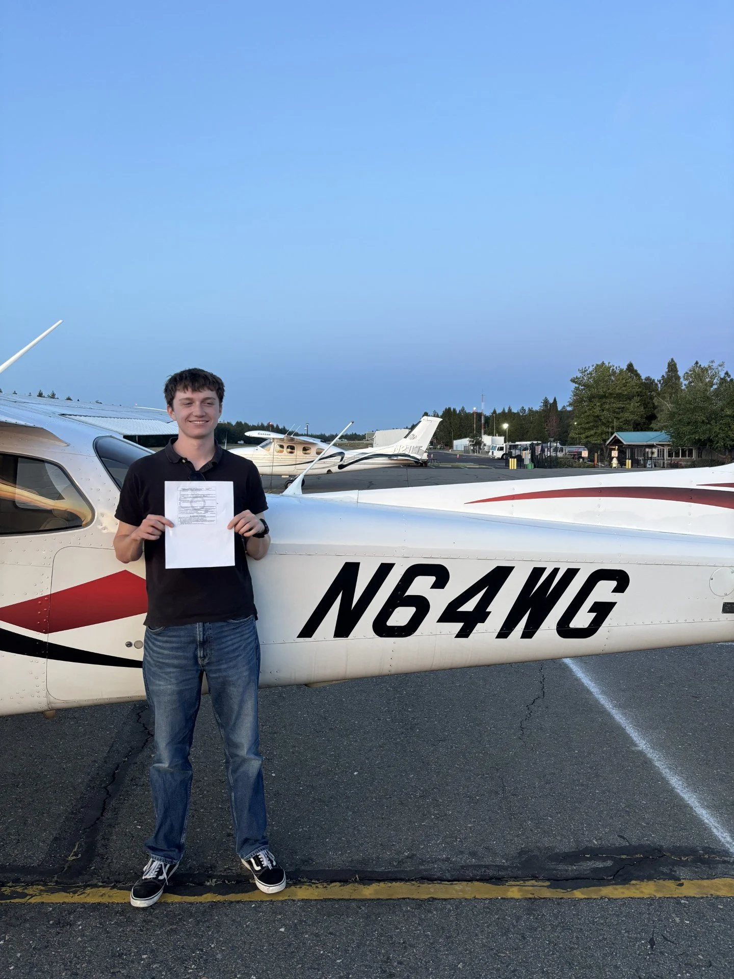 Young man holding a paper standing next to a small white airplane at an airport during twilight.