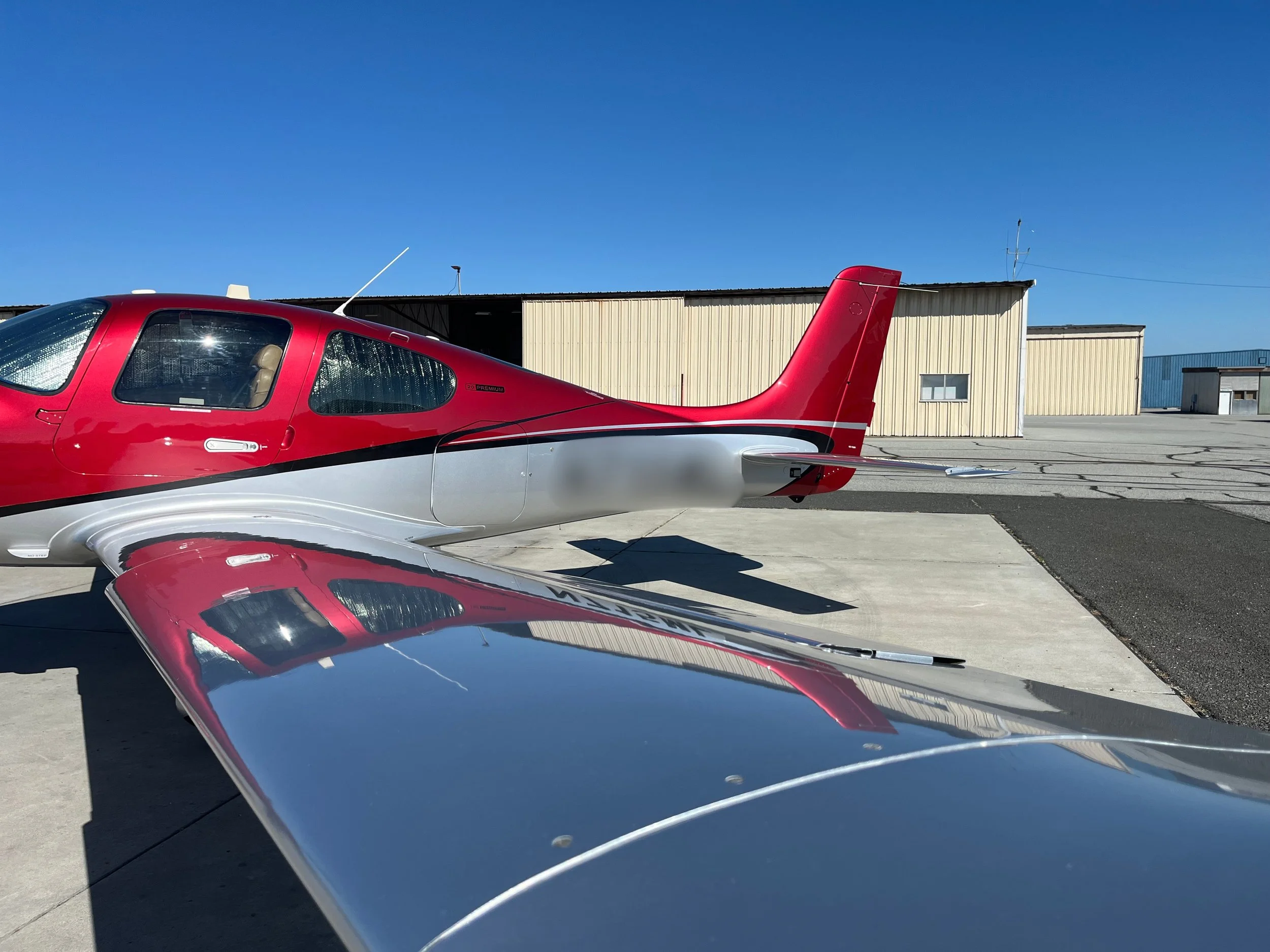 A red and silver small aircraft parked on a tarmac in front of a hangar under a clear blue sky.