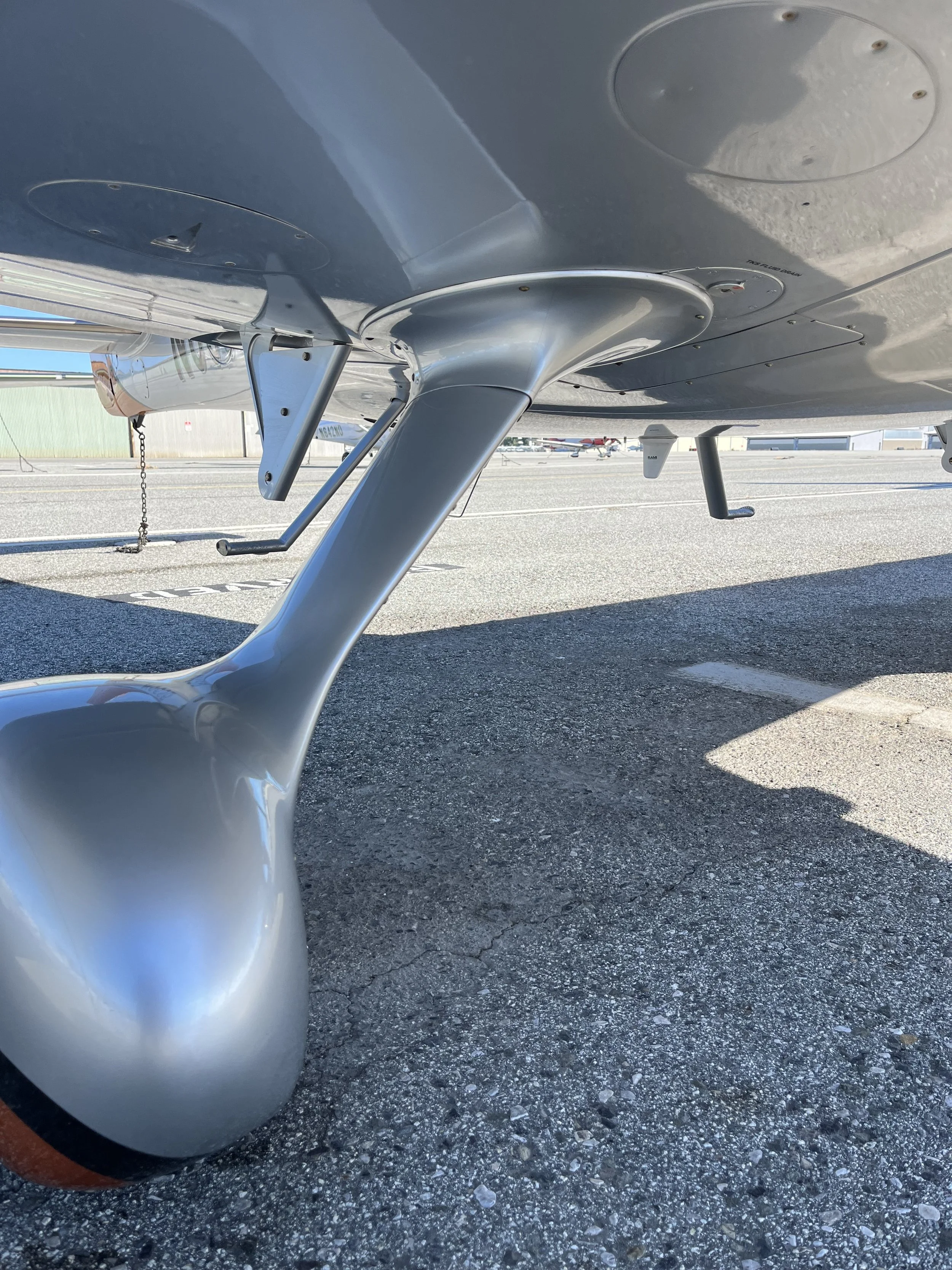 Close-up view of the underside of a small aircraft aircraft, showing the landing gear and part of the fuselage on an asphalt surface at an airport.