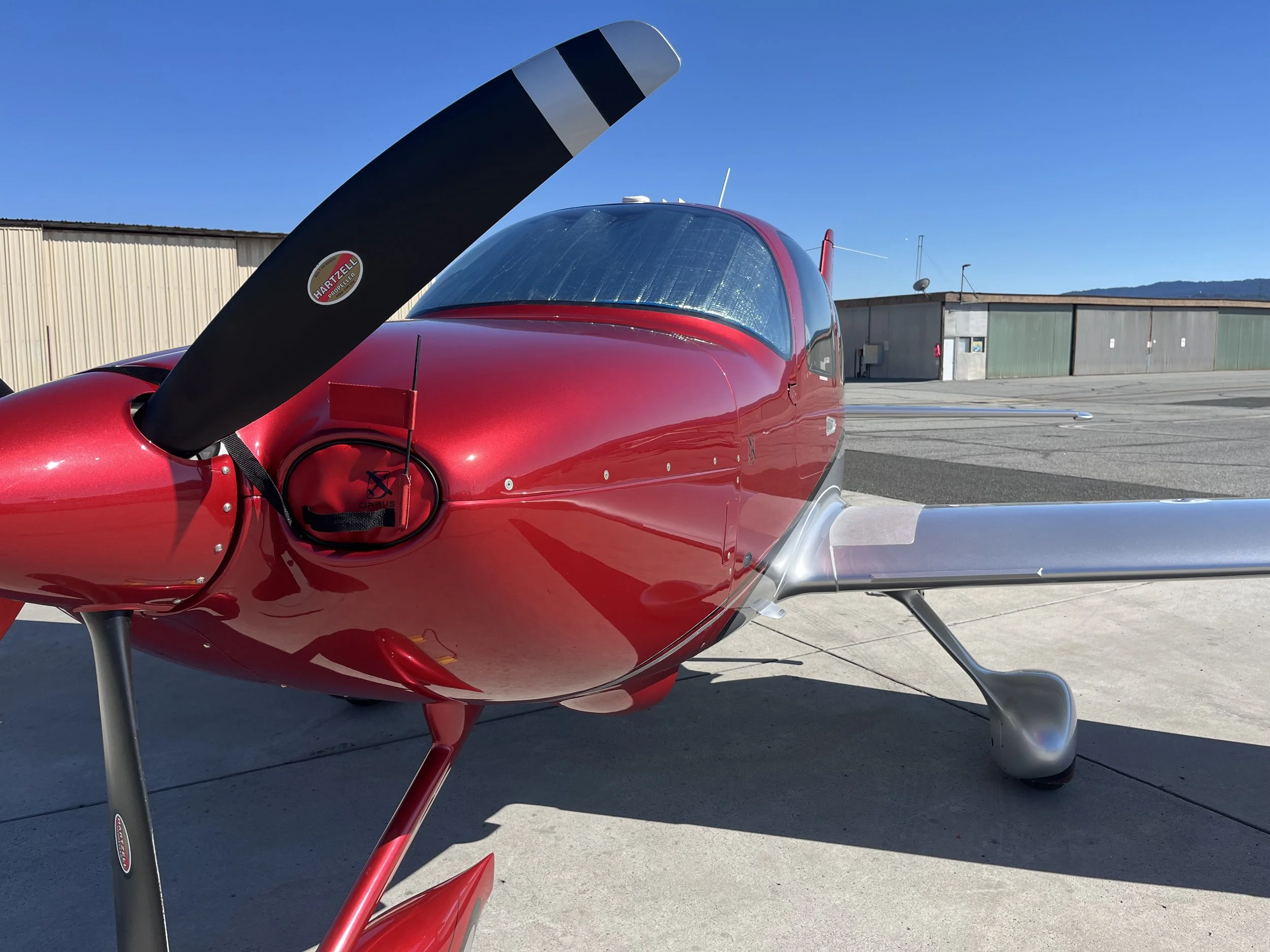 A small red propeller airplane parked on a tarmac with a beige hangar and blue sky in the background.