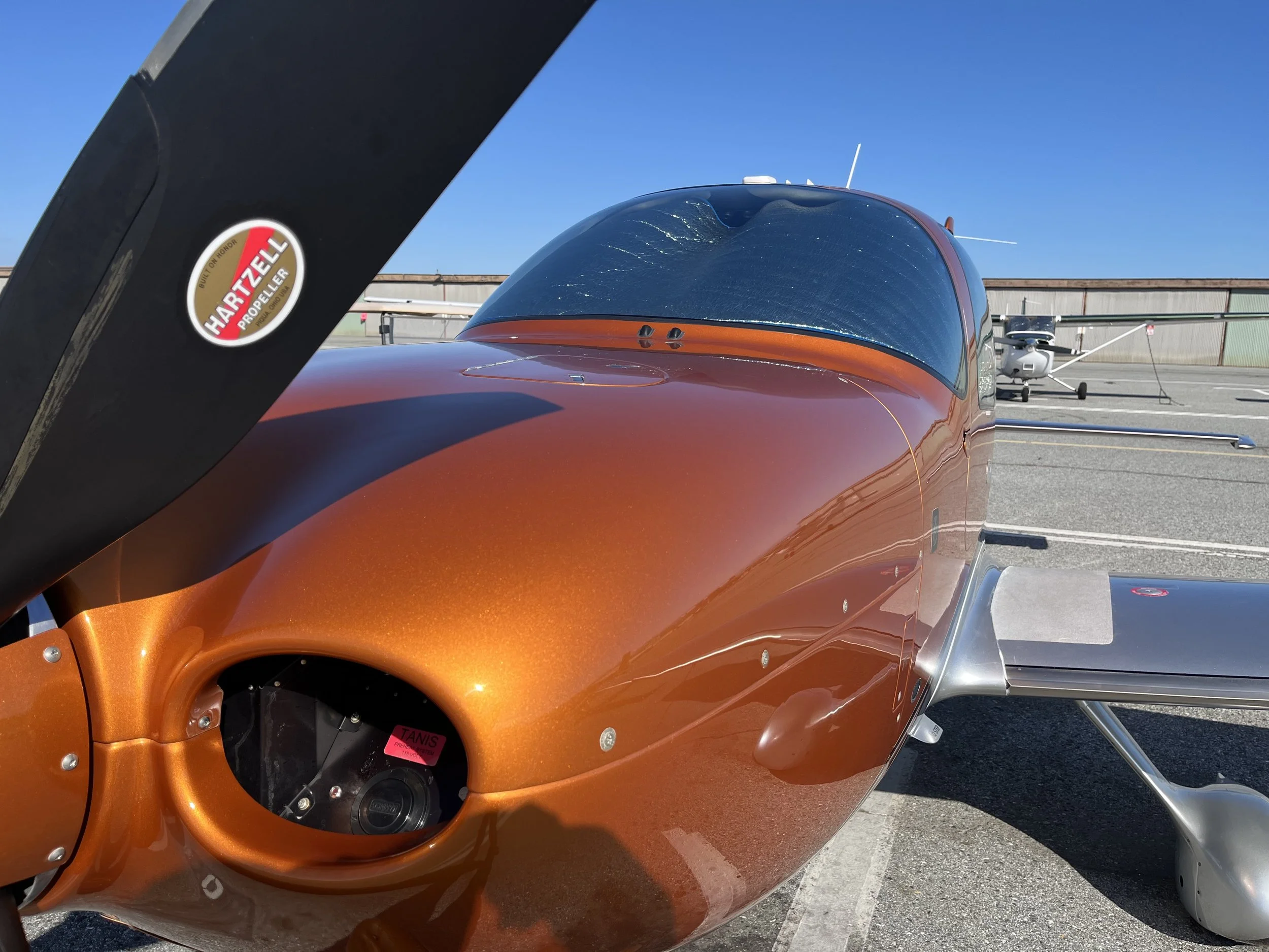 Close-up of a bright orange aircraft cockpit with an empty instrument panel and a large tinted windshield. Part of another aircraft in the background, against a clear blue sky.