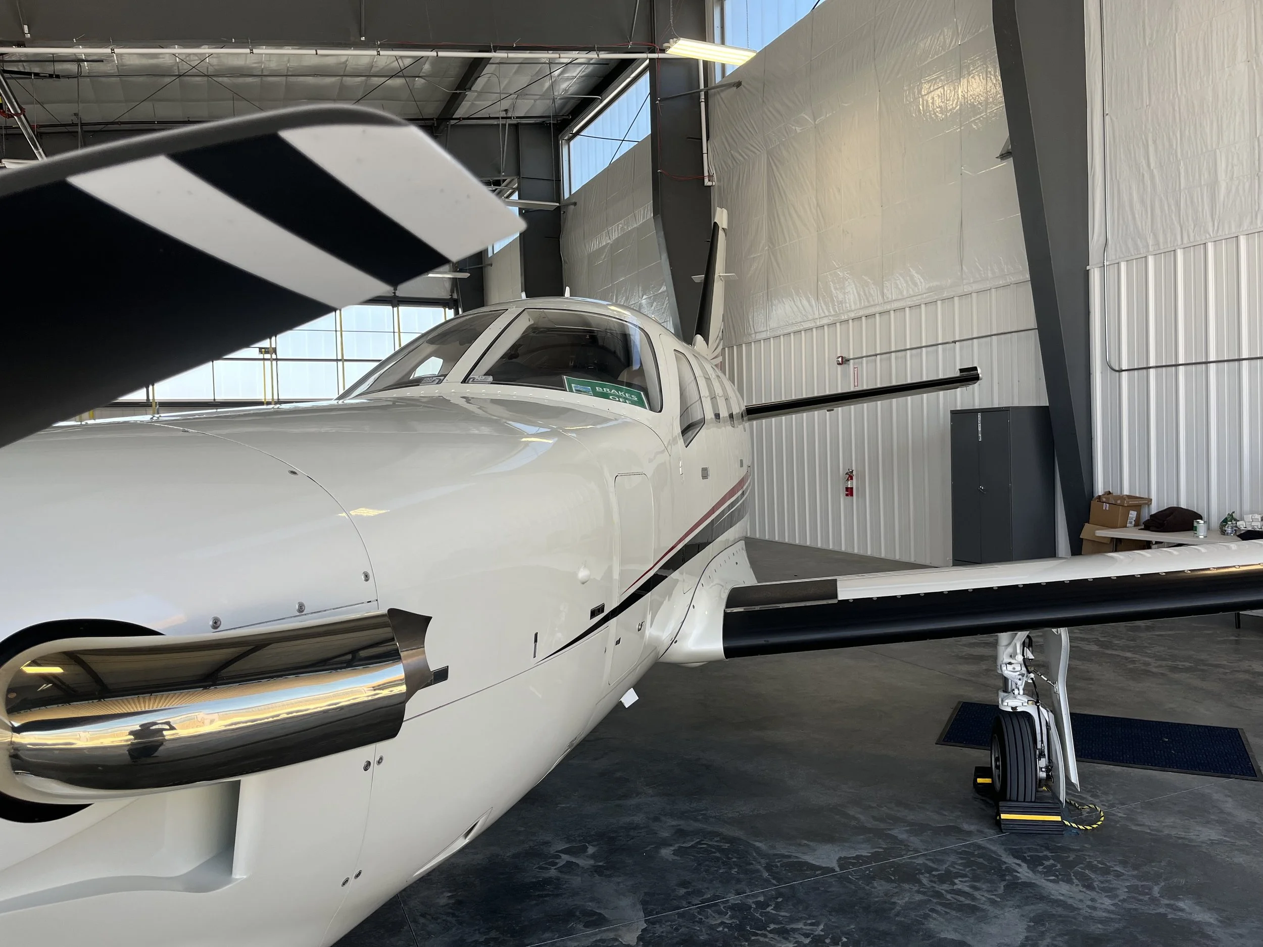 A white aircraft with black and red accents is inside a large hangar. The cockpit canopy is closed, and the aircraft's nose and part of its wings are visible. The hangar has a high ceiling with metal framing, large windows, and a wall with white pane
