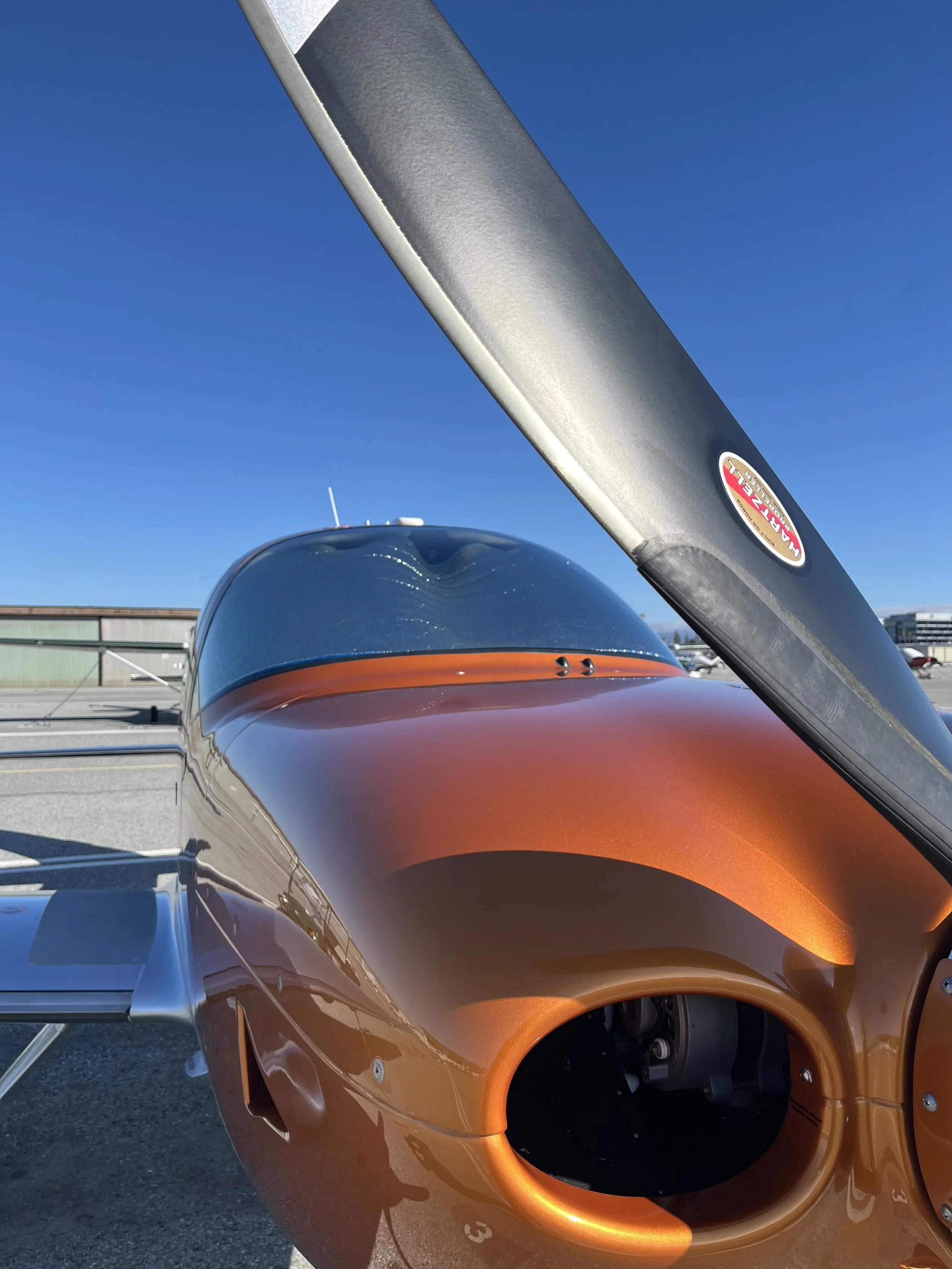 Close-up of a racing car with an orange body, showing the front wheel well and a part of the windshield, with a metallic bar crossing in front and a clear blue sky in the background.
