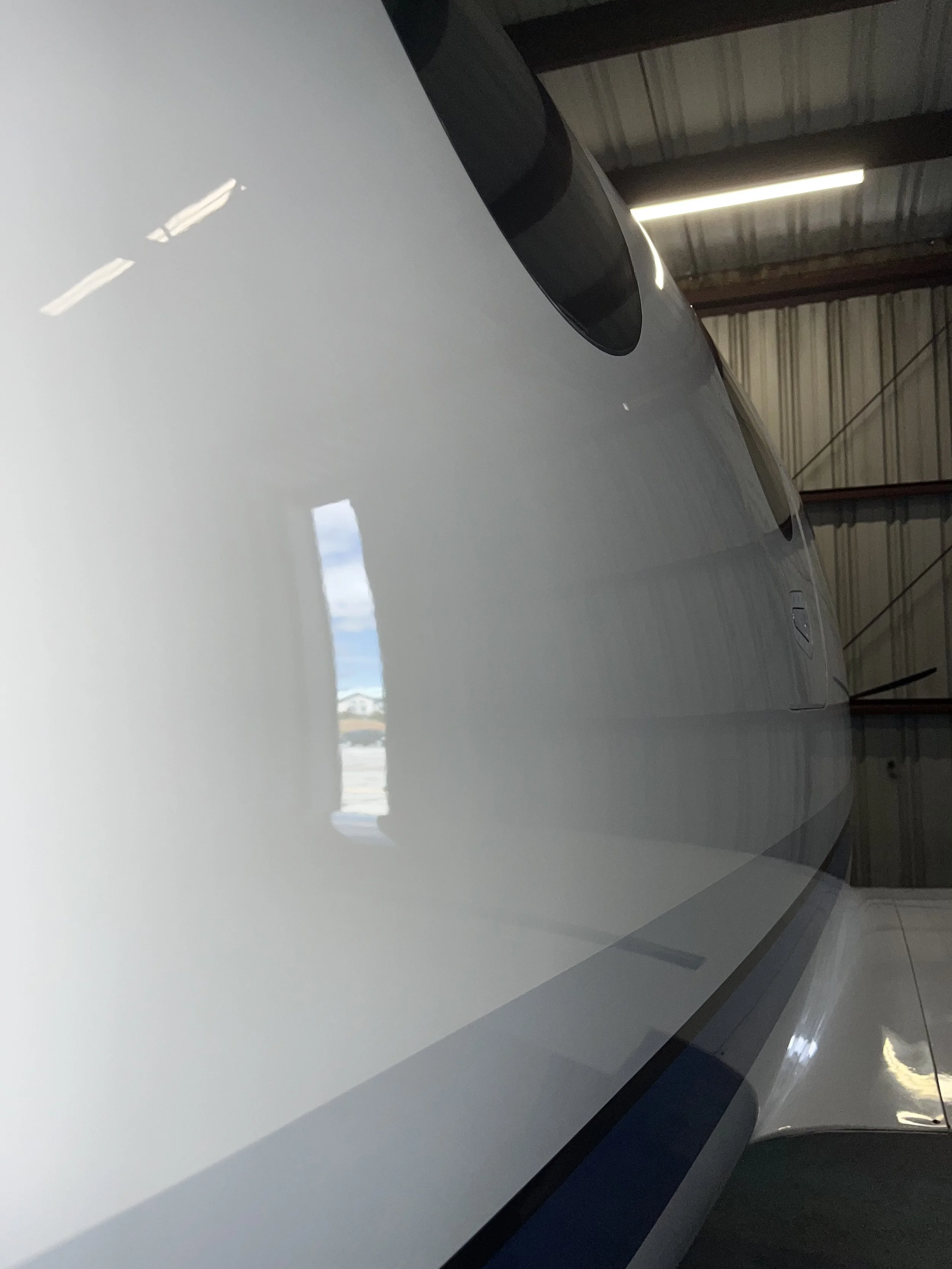 Close-up of a white airplane fuselage, showing a dark tinted window, inside a hangar with a metal roof and partial view of the sky.