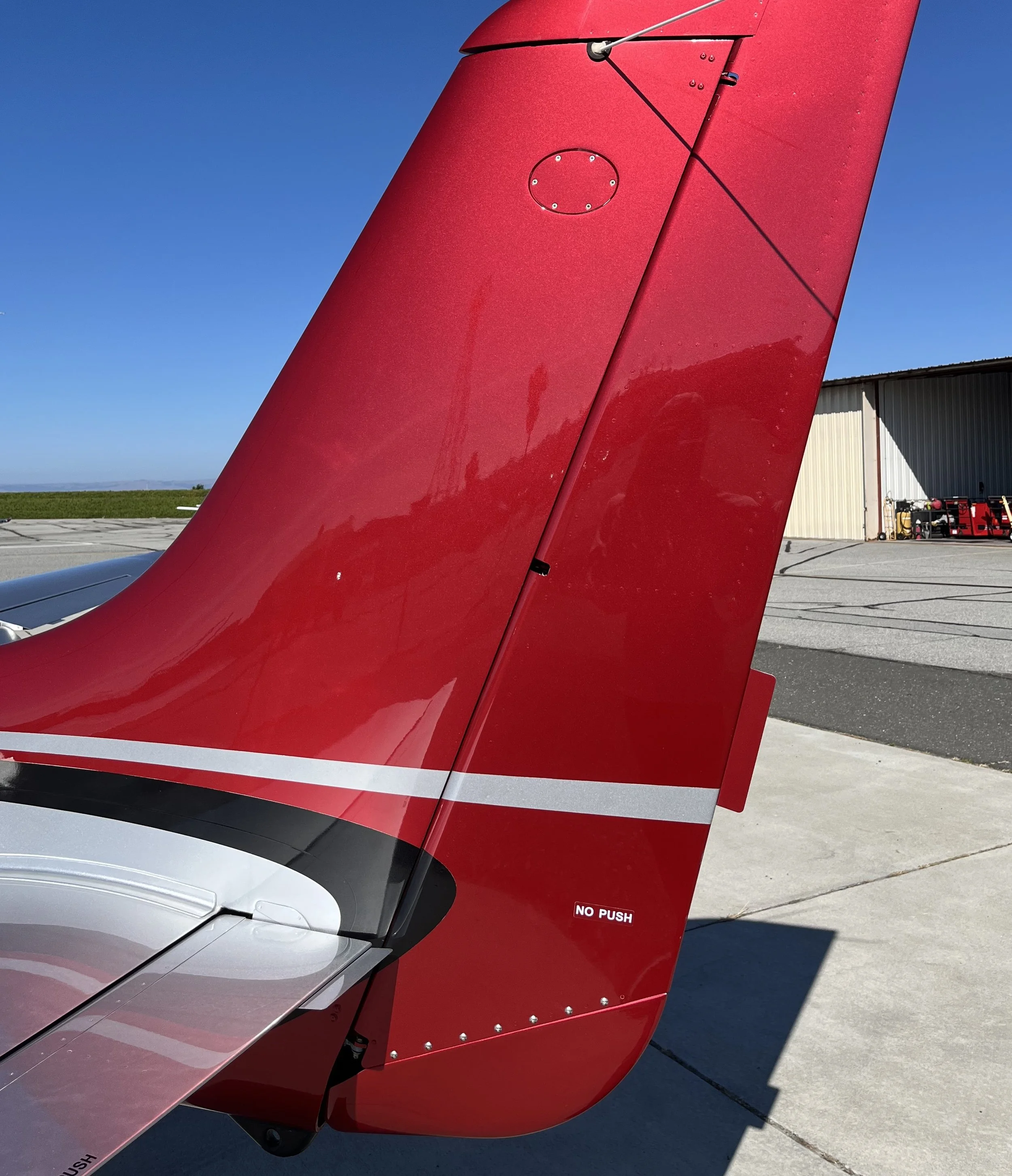 Close-up of the tail section of a red aircraft with a part of the landing gear visible, and a small sign that says 'NO PUSH' on the fuselage, parked on a tarmac under a clear blue sky with hangar and equipment in the background.