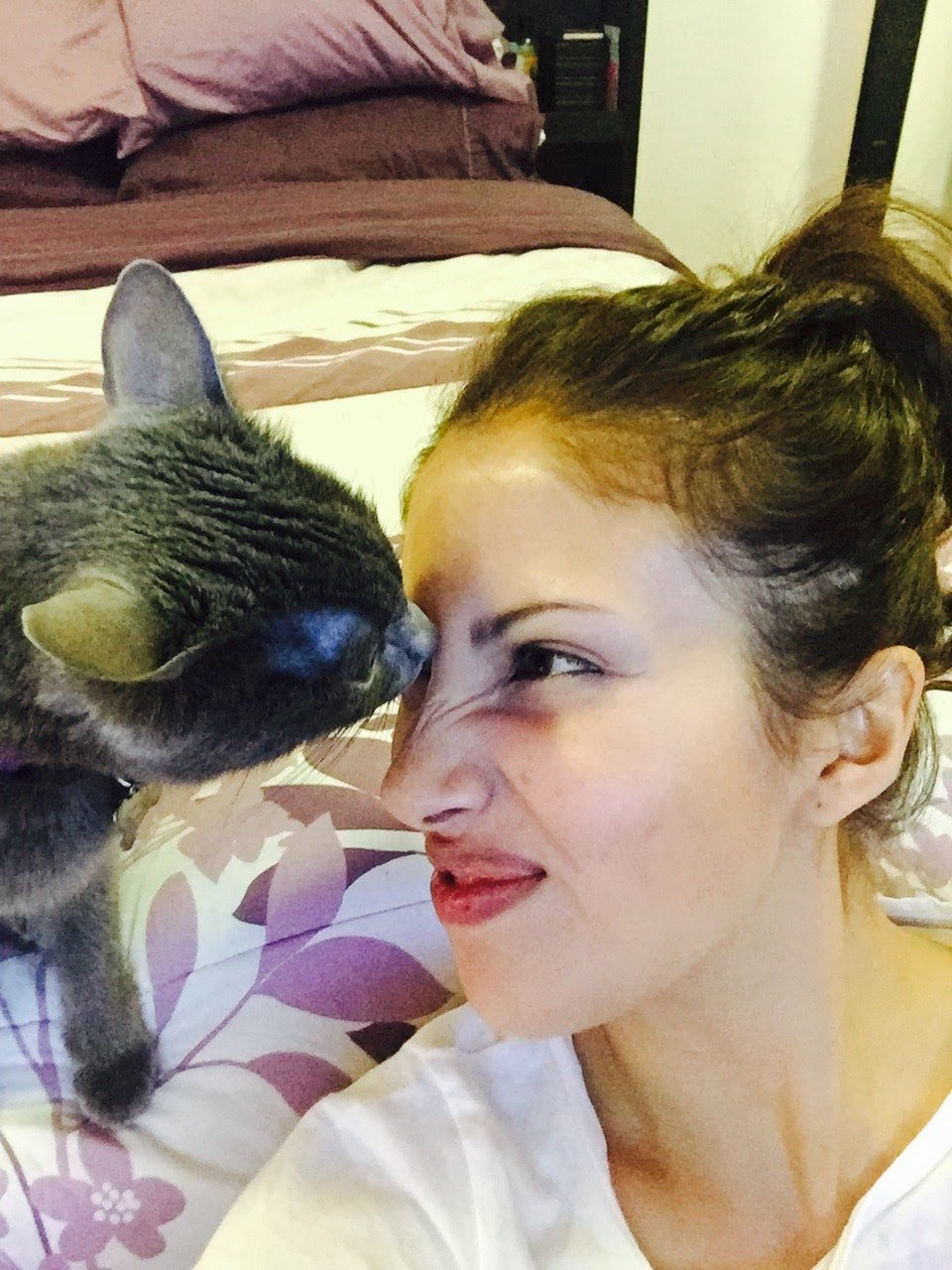 Mujer sonriendo cerca de un gato gris que se acerca a su cara en una cama con ropa de cama morada y blanca.