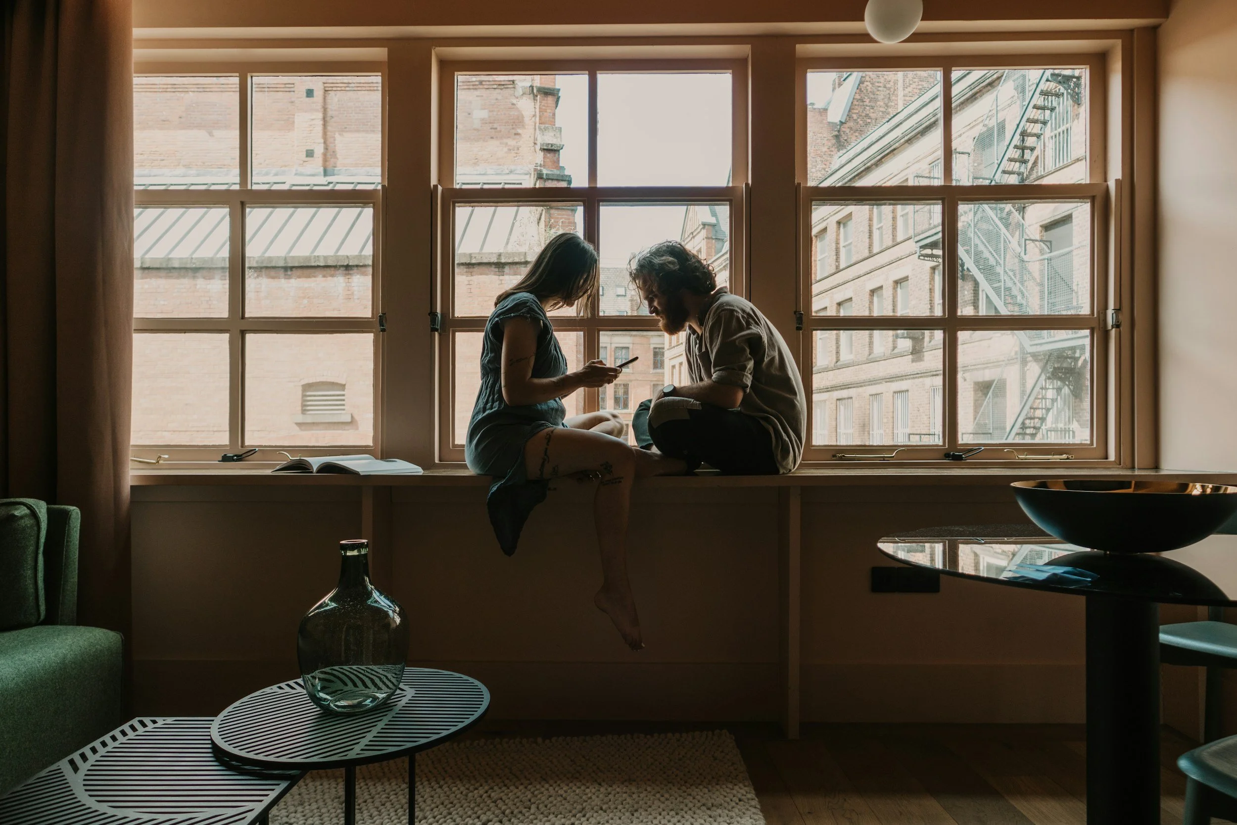 Two people sitting on a window ledge, looking at a phone inside an apartment with large windows showing brick buildings outside.