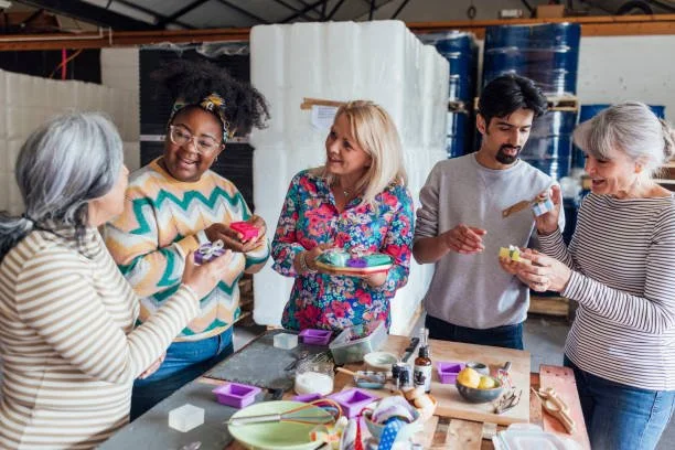 Group of five diverse women smiling and chatting while decorating soap bars in a workshop setting.