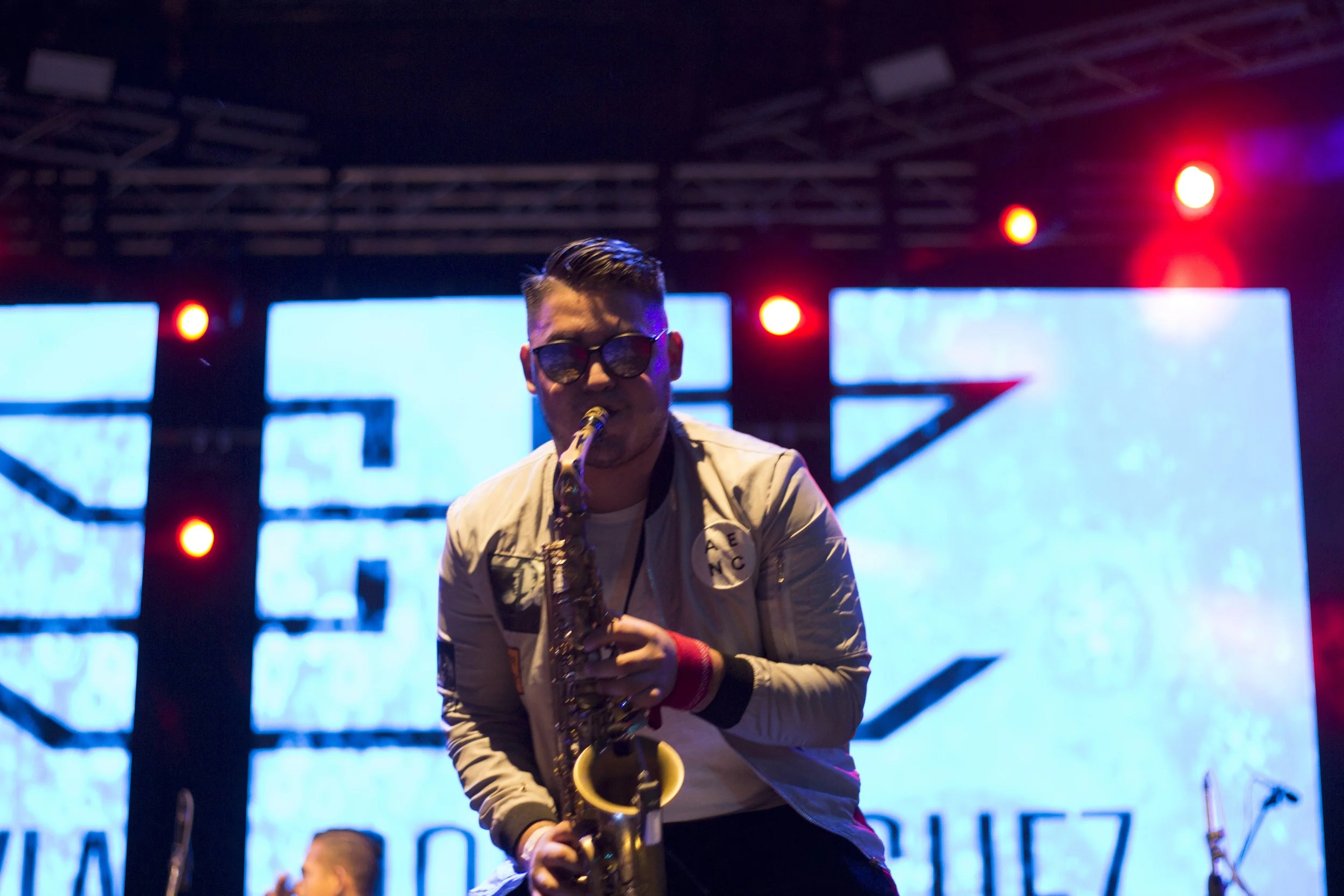 A musician wearing sunglasses and a light-colored jacket plays a saxophone on stage with blue screen backgrounds and red stage lights.